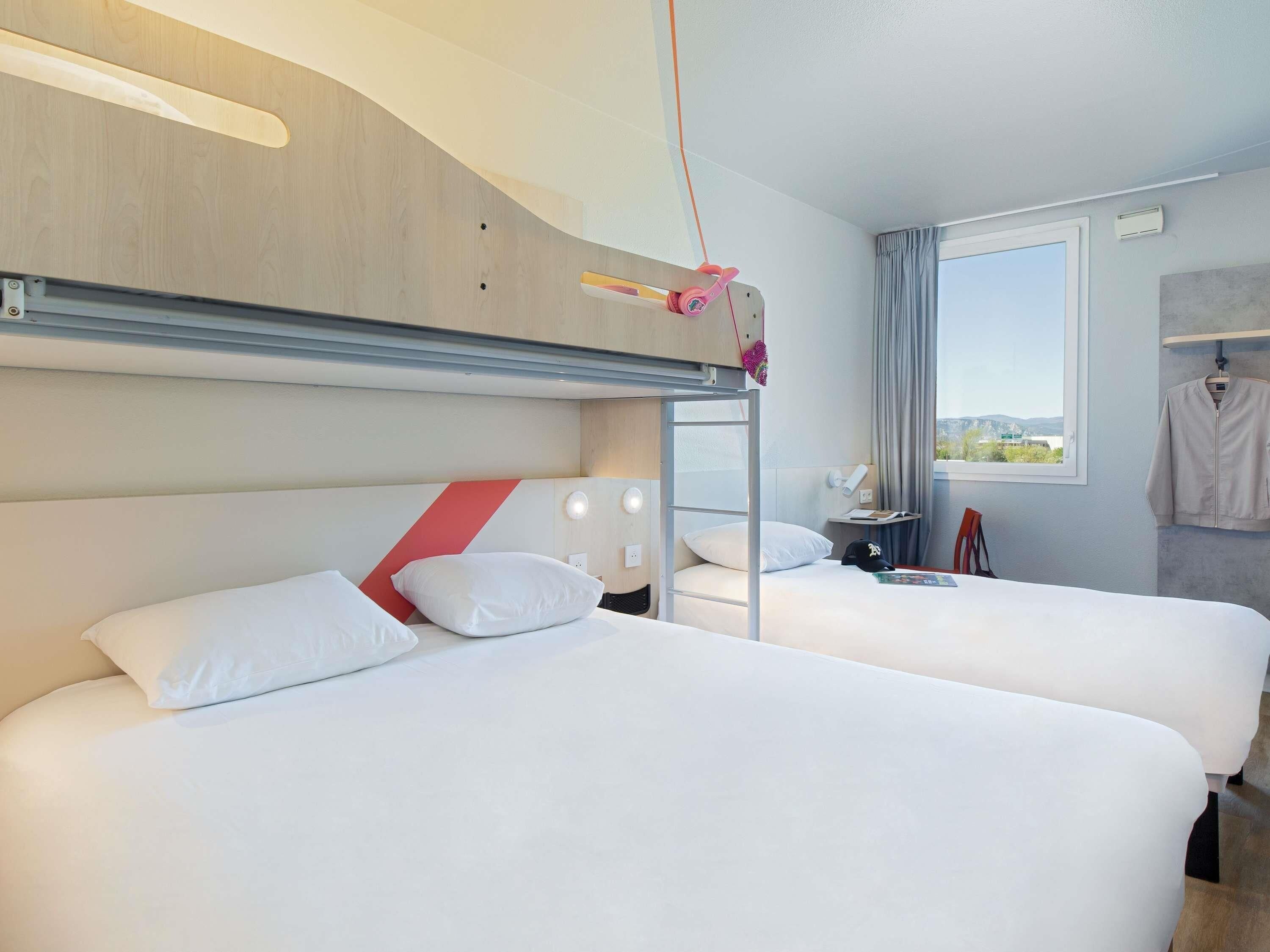 A hotel room with two beds, a bunk bed, a window with a view, and a hanging coat rack.