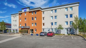 A multi-story hotel with a sign reading 'ibis budget,' a parking lot with several cars, and a clear blue sky. - ibis budget Valence Nord Plateau des Couleures (Saint-Marcel-les-Valence)