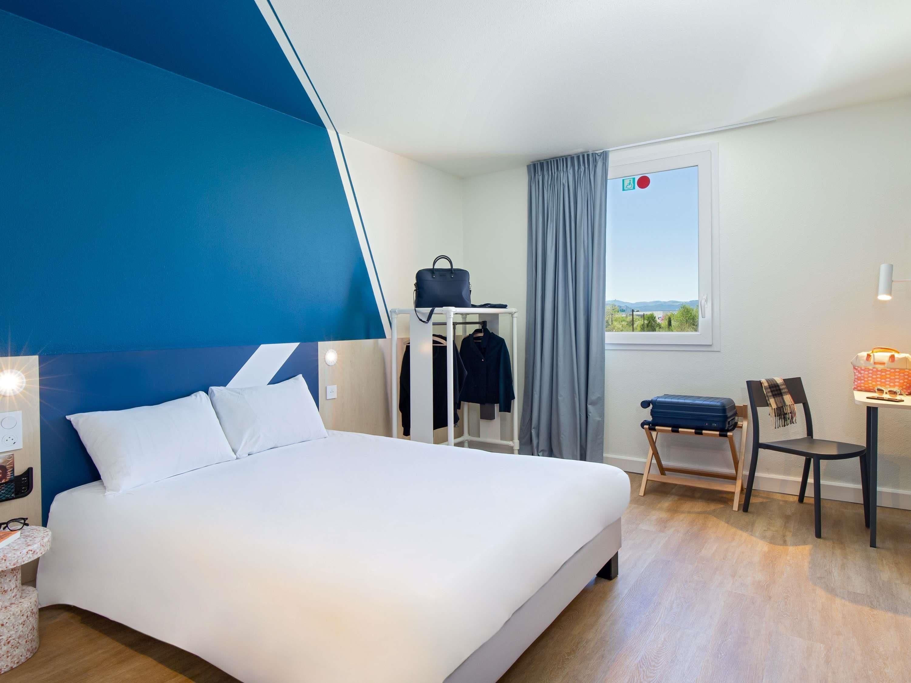 Una camera d'albergo con un letto, una sedia, un tavolo, un appendiabiti e una finestra con vista.