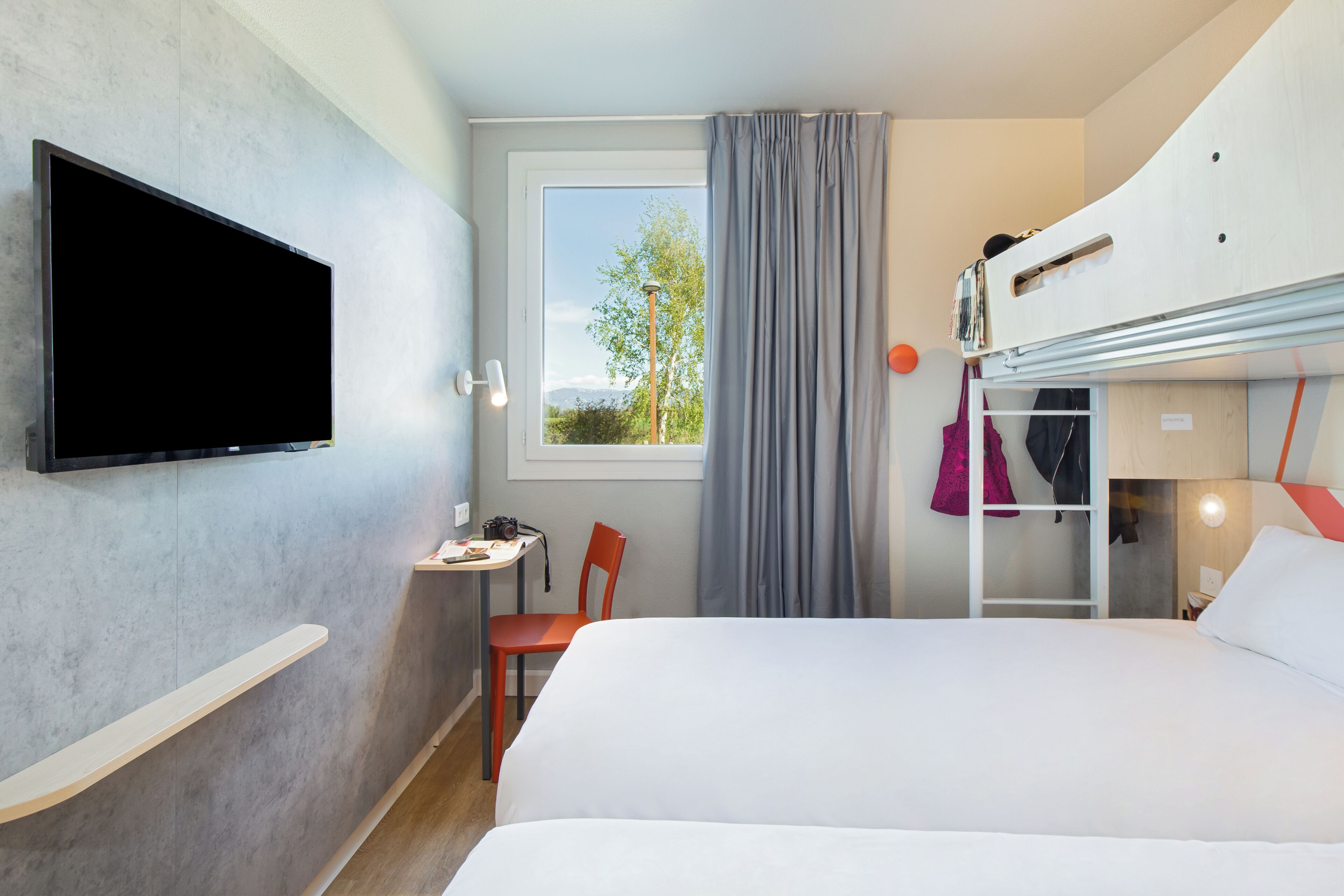 Une chambre d’hôtel avec un lit, un téléviseur fixé au mur, un petit bureau avec une chaise, une fenêtre avec des rideaux et un lit superposé.