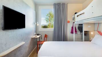 Une chambre d’hôtel avec un lit, un téléviseur fixé au mur, un petit bureau avec une chaise, une fenêtre avec des rideaux et un lit superposé.