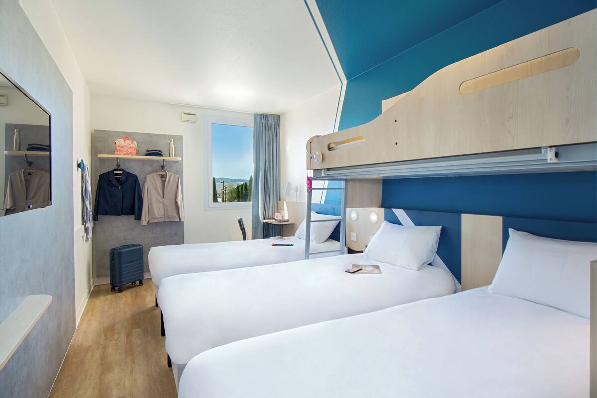 A hotel room with bunk beds, a suitcase, a mirror, and a window with a view.
