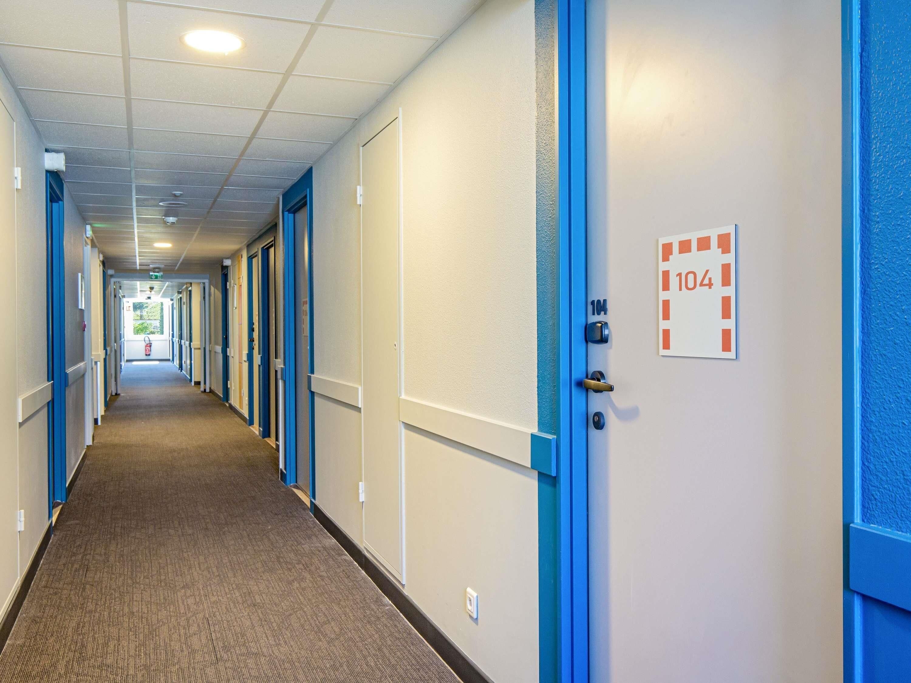 Un couloir d’hôtel avec des portes numérotées, dont la porte 104, un sol recouvert de moquette et des encadrements de porte bleus.