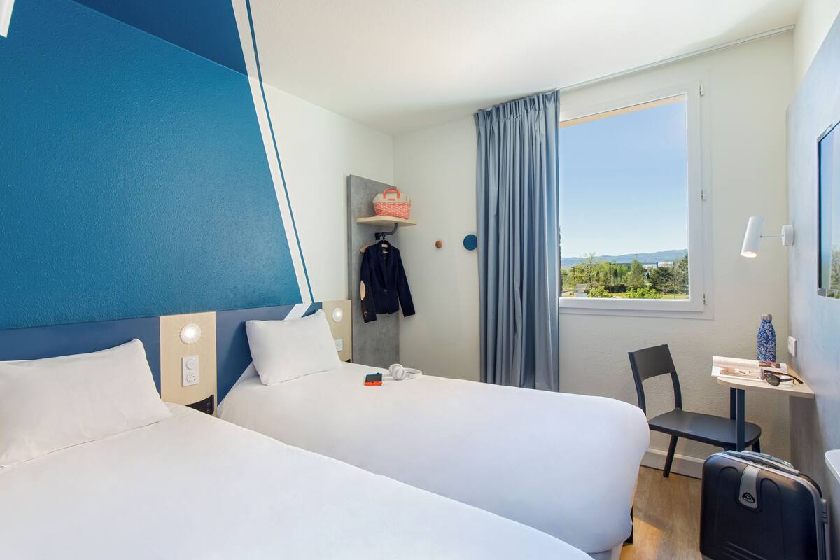 A hotel room with two beds, a desk with a chair, a window with a view, and a small table with a bottle.