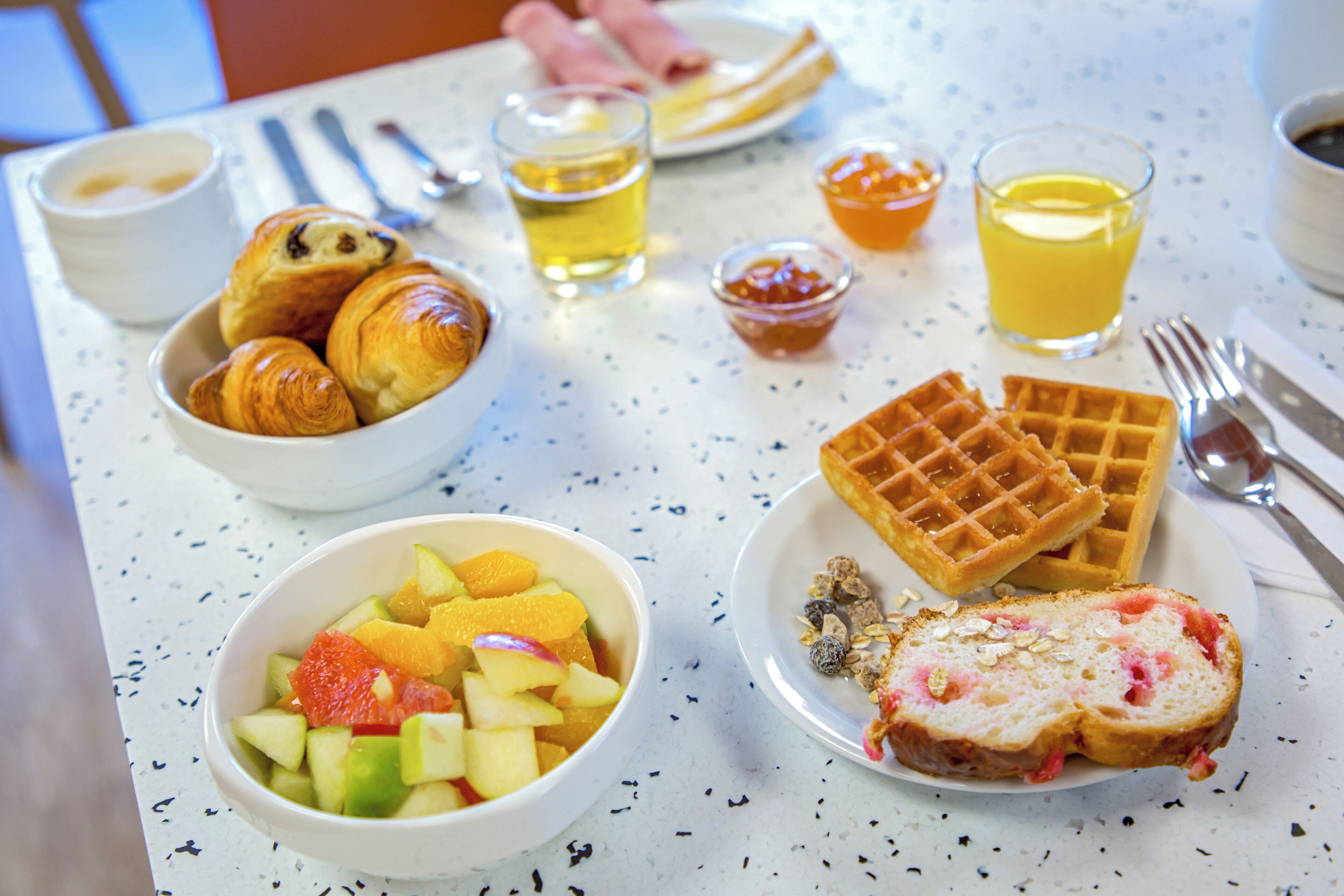Una mesa de desayuno con cruasanes, gofres, ensalada de frutas y una rebanada de pan con mermelada.