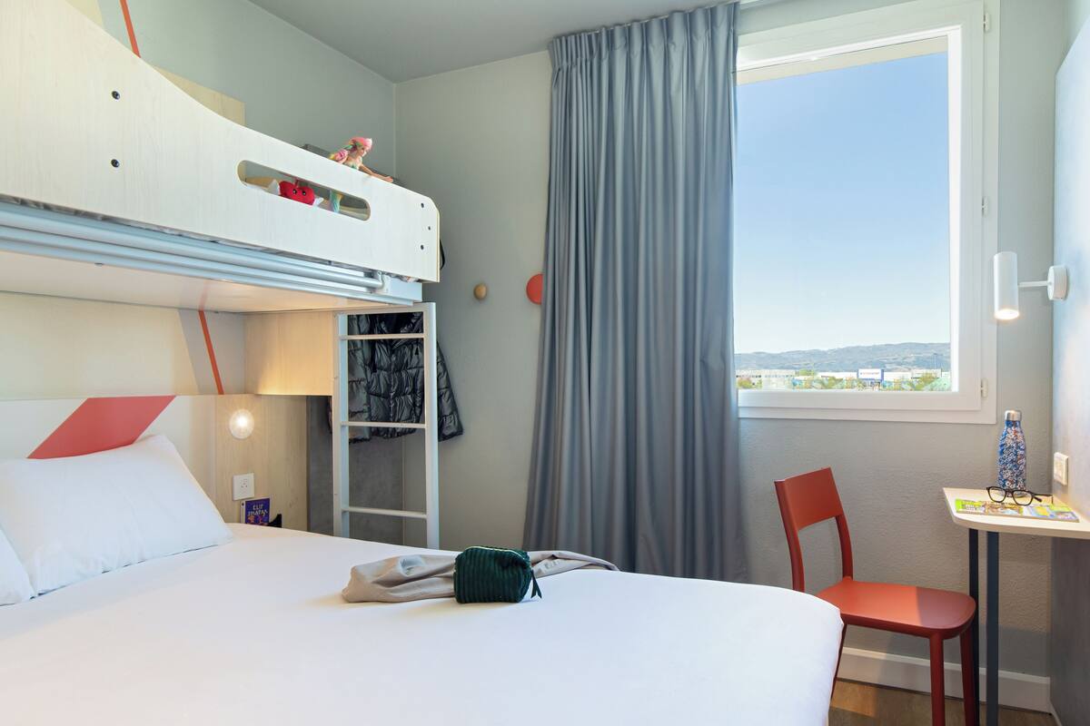 A hotel room with a bunk bed, a desk with a chair, a window with curtains, and a view of the outside.