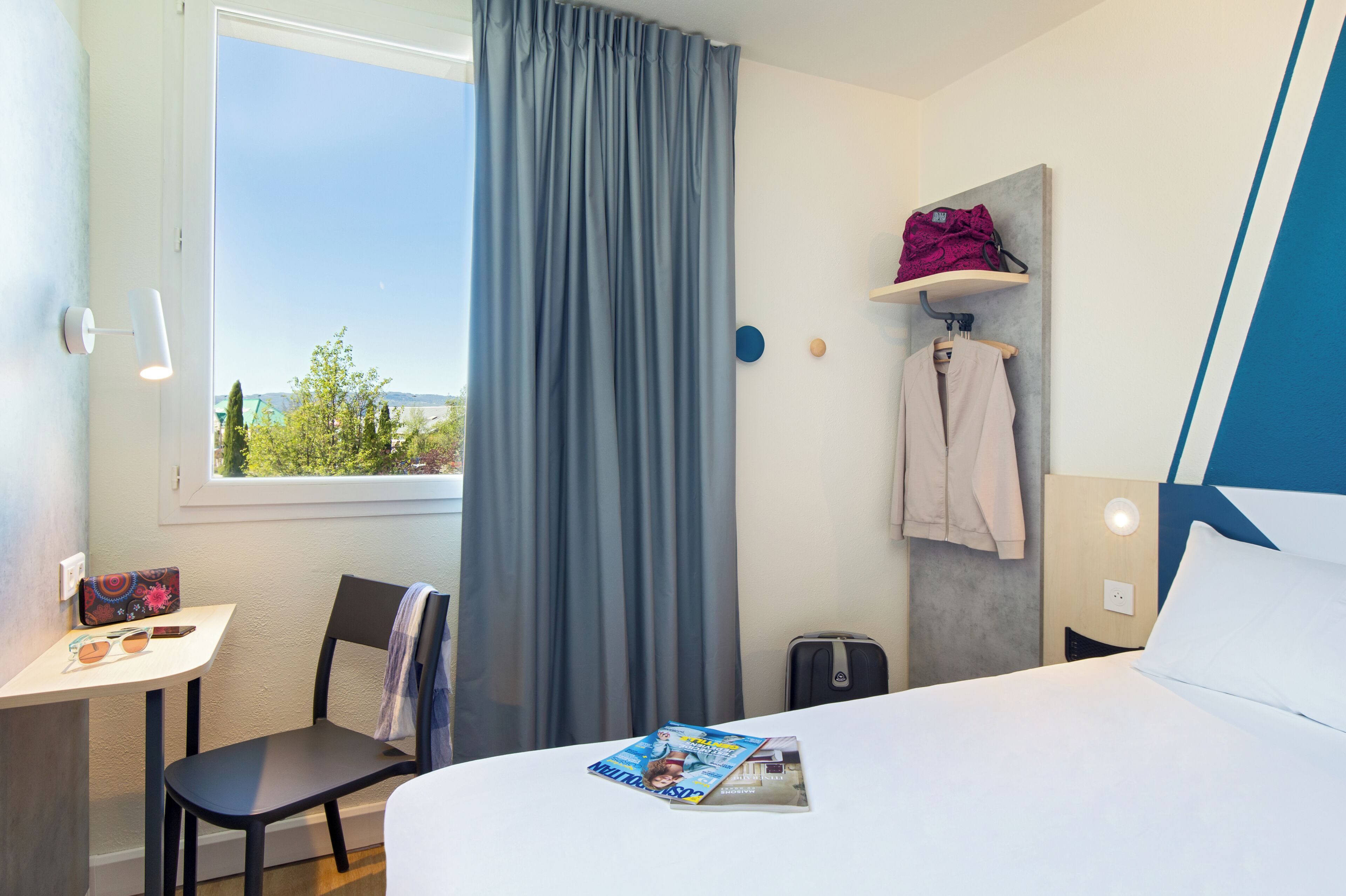 Une chambre d’hôtel avec un lit, une table avec un magazine, une chaise, une fenêtre avec des rideaux, un porte-manteau et une valise.