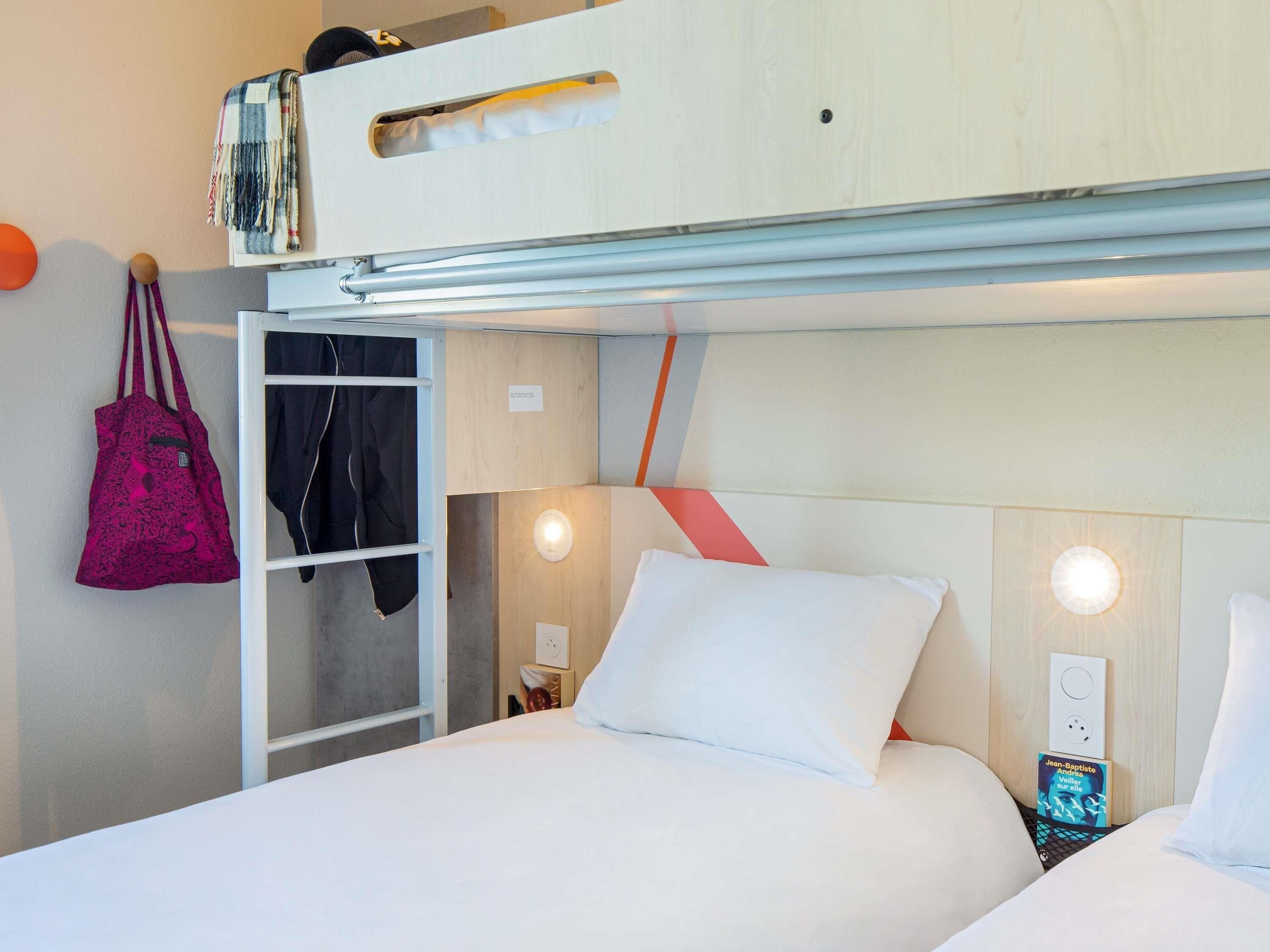 Una habitación de hotel con literas, un armario, una mesita de noche con lámpara y un enchufe en la pared.