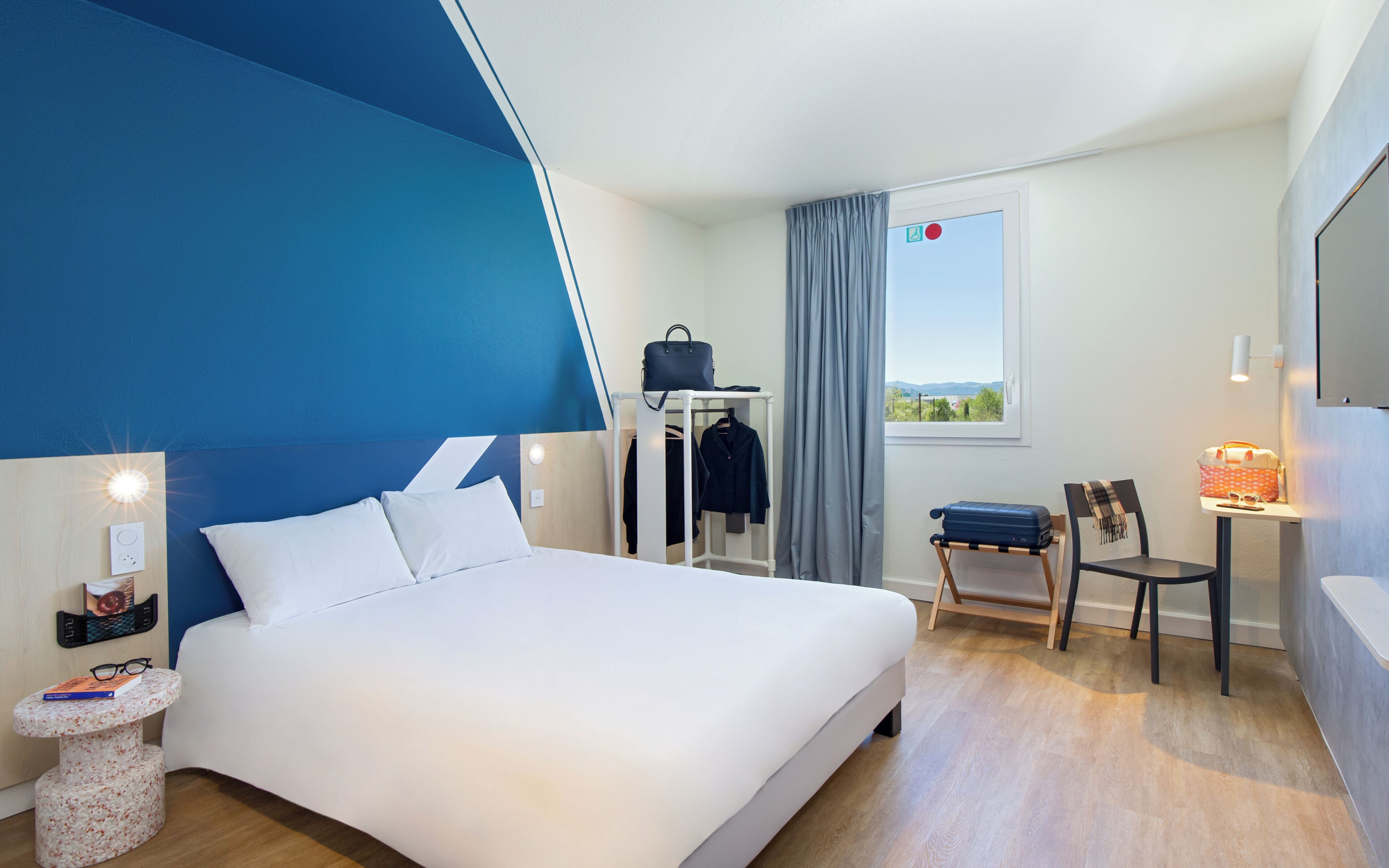 Quarto de hotel com cama, escrivaninha, cadeira, guarda-roupa e janela com vista.
