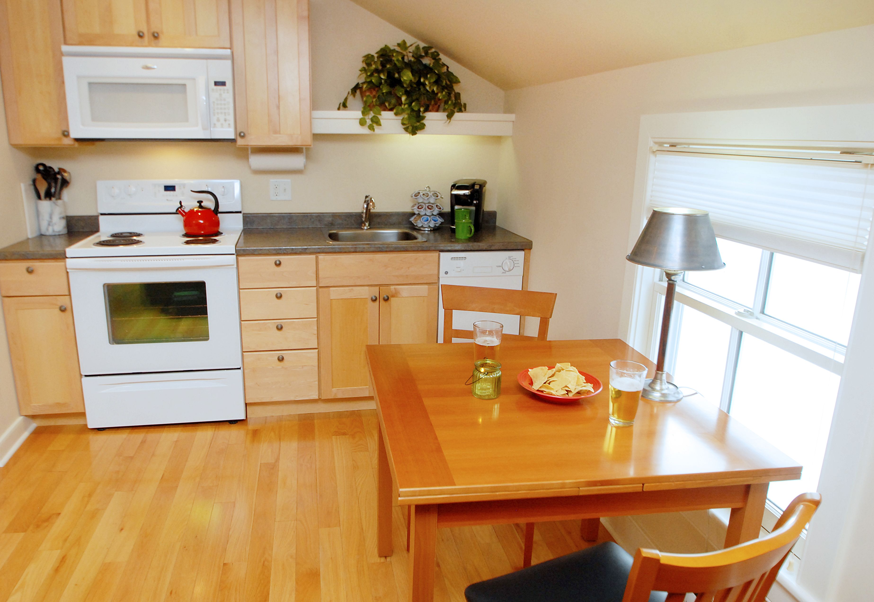 A compact kitchen with a microwave, stove, and sink. There's a dining table with chairs and a lamp.