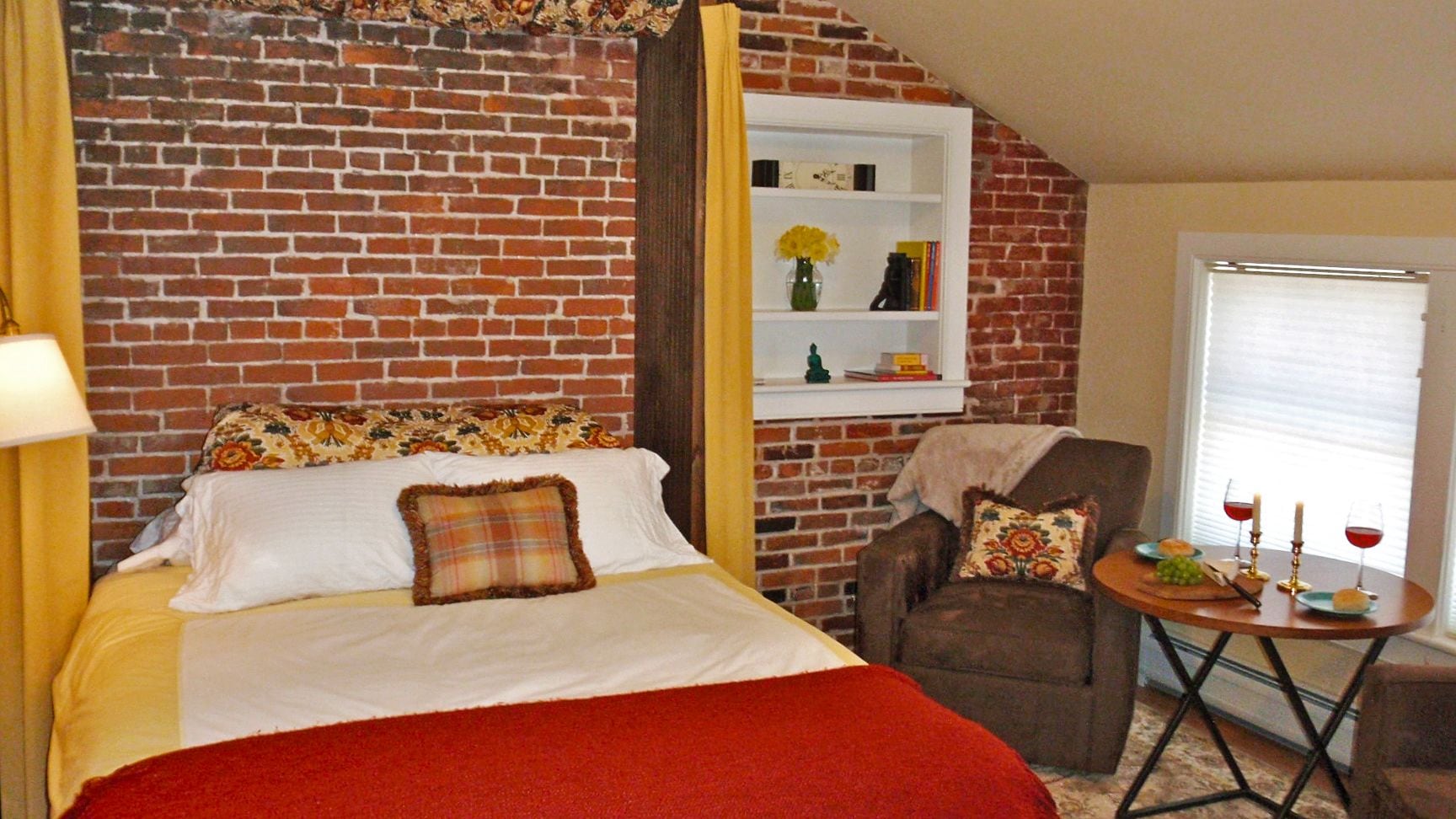 A room with a brick wall, a bed with white bedding, a chair, a small table with a candle and glass, and a window with blinds.