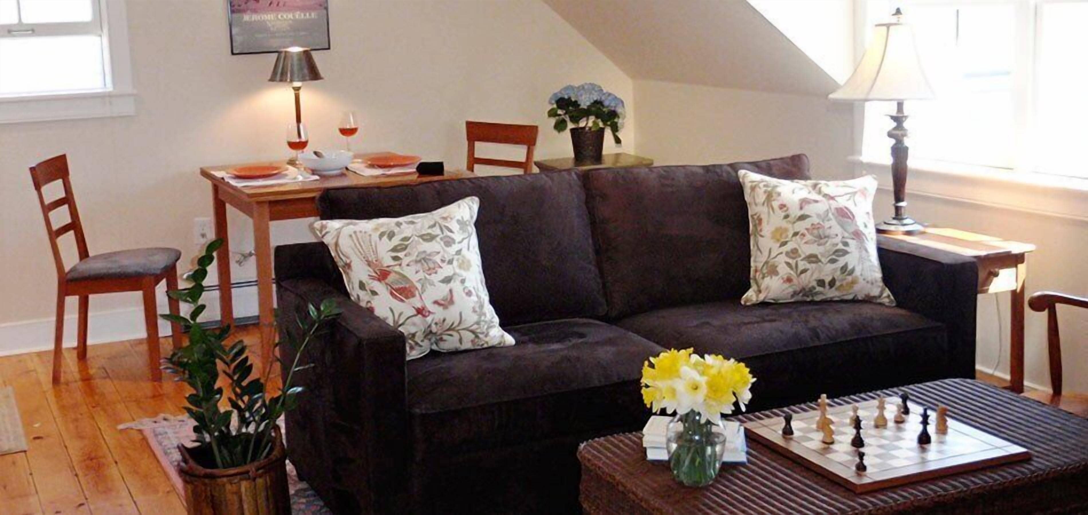 A living room with a dark brown sofa, two floral cushions, a chessboard, and a vase with yellow flowers.