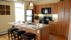 A kitchen with wooden cabinets, a stainless steel refrigerator, a microwave, and a stove. There are three bar stools and a sink with a faucet. - Butler House (Stowe)