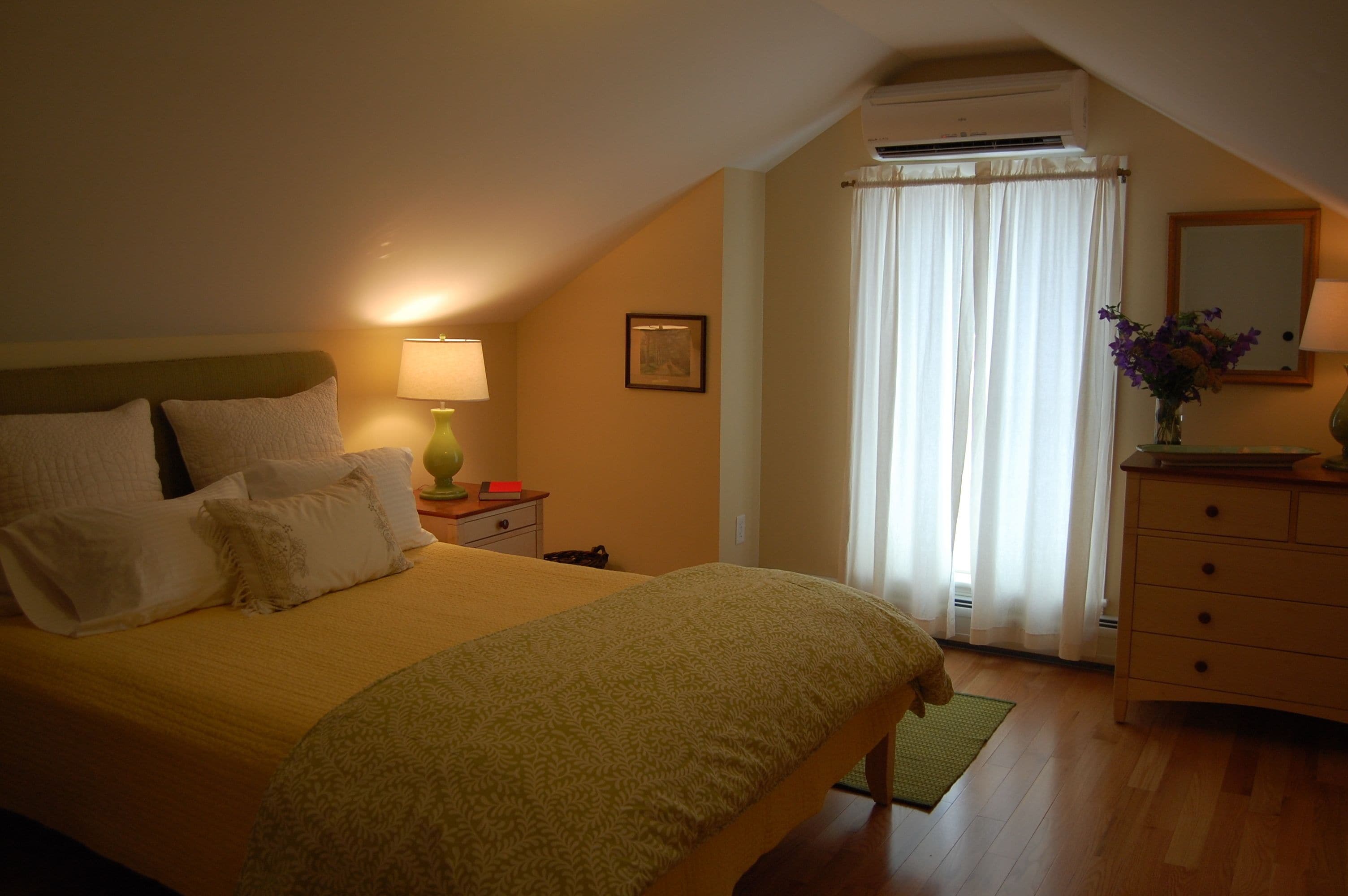 A bedroom with a bed, bedside table, lamp, dresser, and a window with curtains.