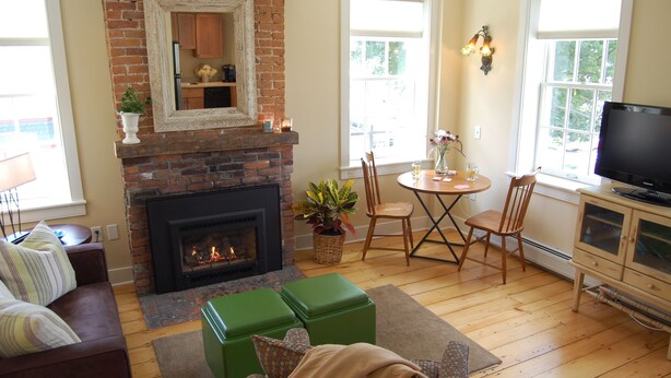 A cozy living room with a brick fireplace, green ottomans, a TV, and a dining area with chairs and a table. - Butler House (Stowe)