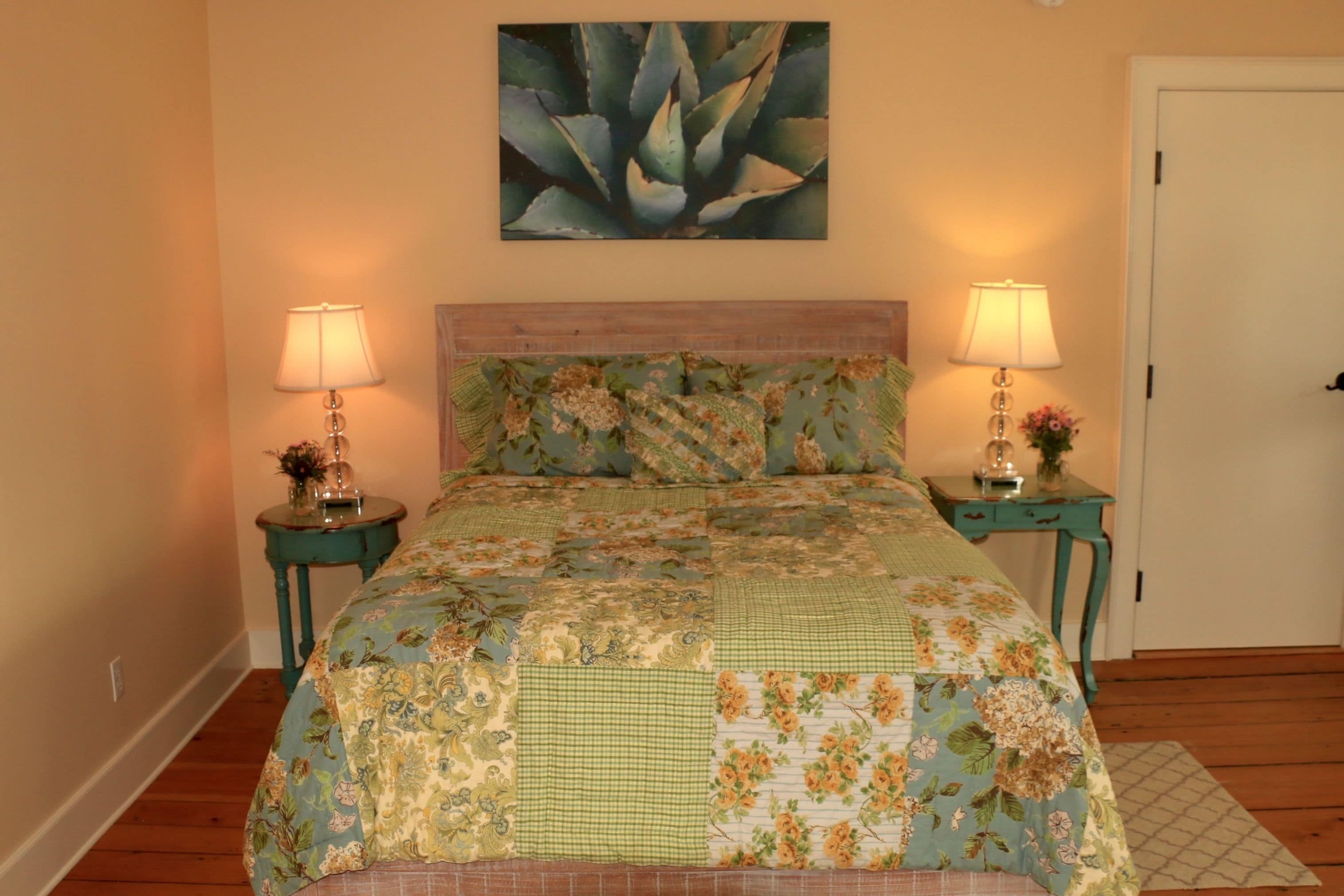 A bedroom with a floral bedspread, two bedside tables with lamps, a door, and a wall-mounted painting.