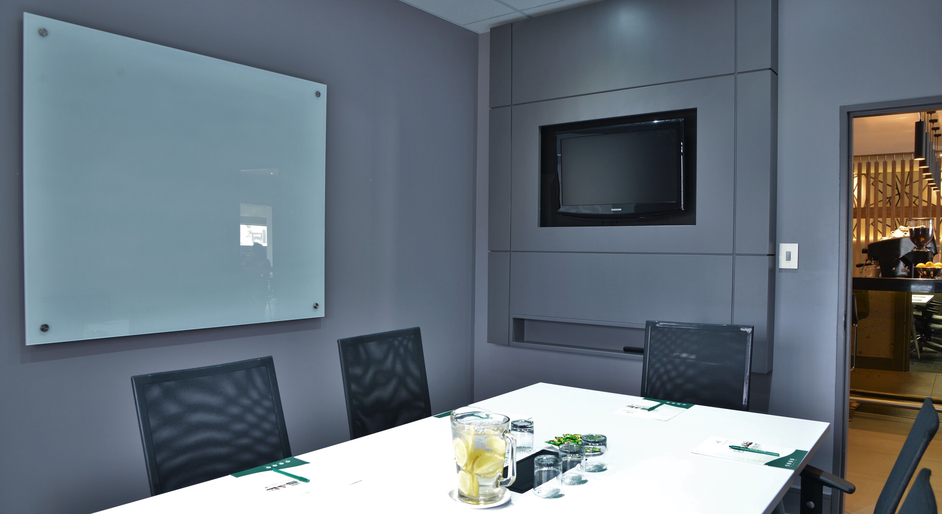Un salón de conferencias con una pizarra grande, un televisor de pantalla plana y una mesa dispuesta con una jarra de agua, vasos y condimentos.