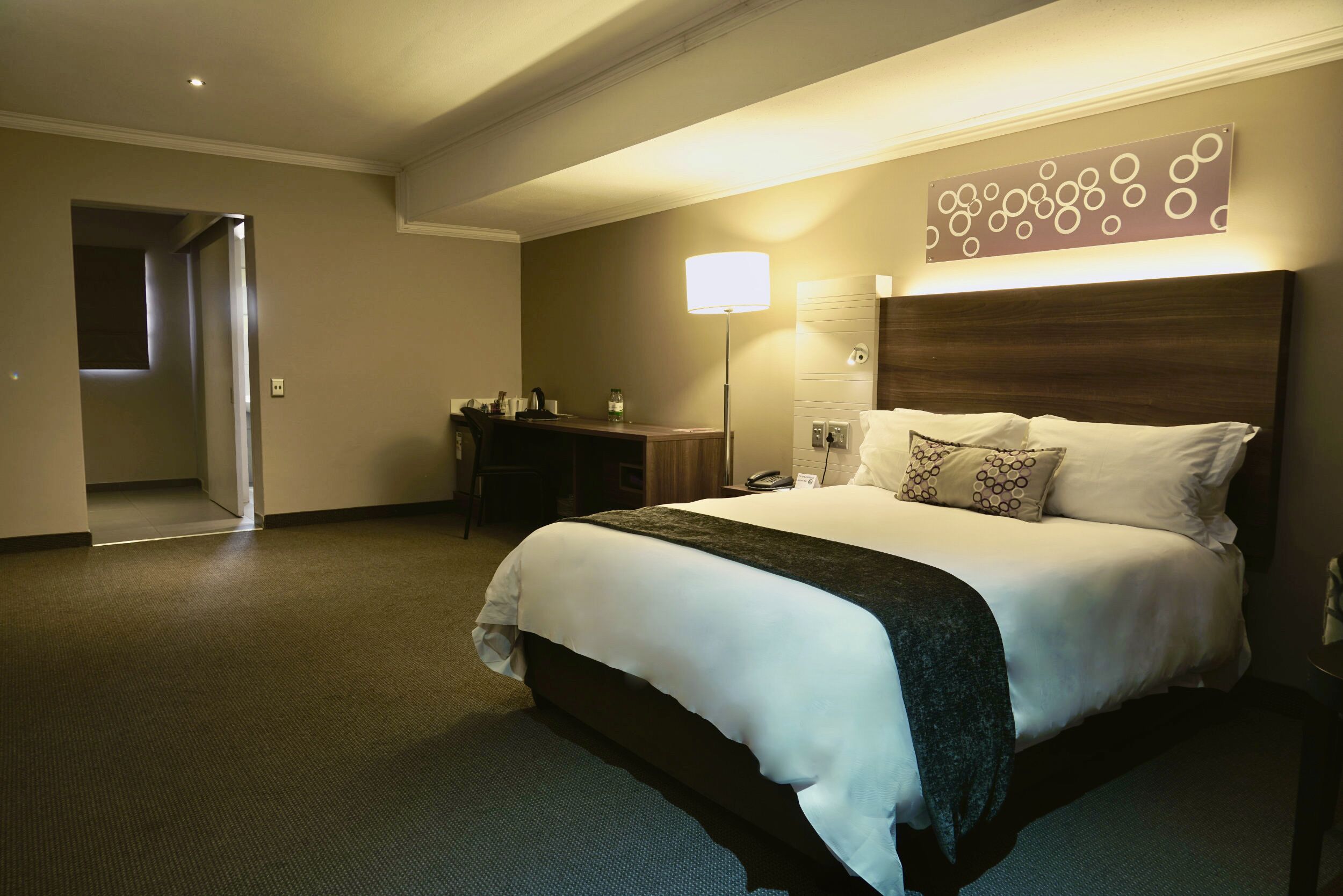 A hotel room with a large bed, a desk, a lamp, and a wall art piece.