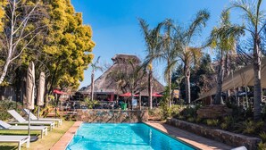 Una piscina con sillas de descanso y sombrillas, rodeada de palmeras y una formación rocosa.