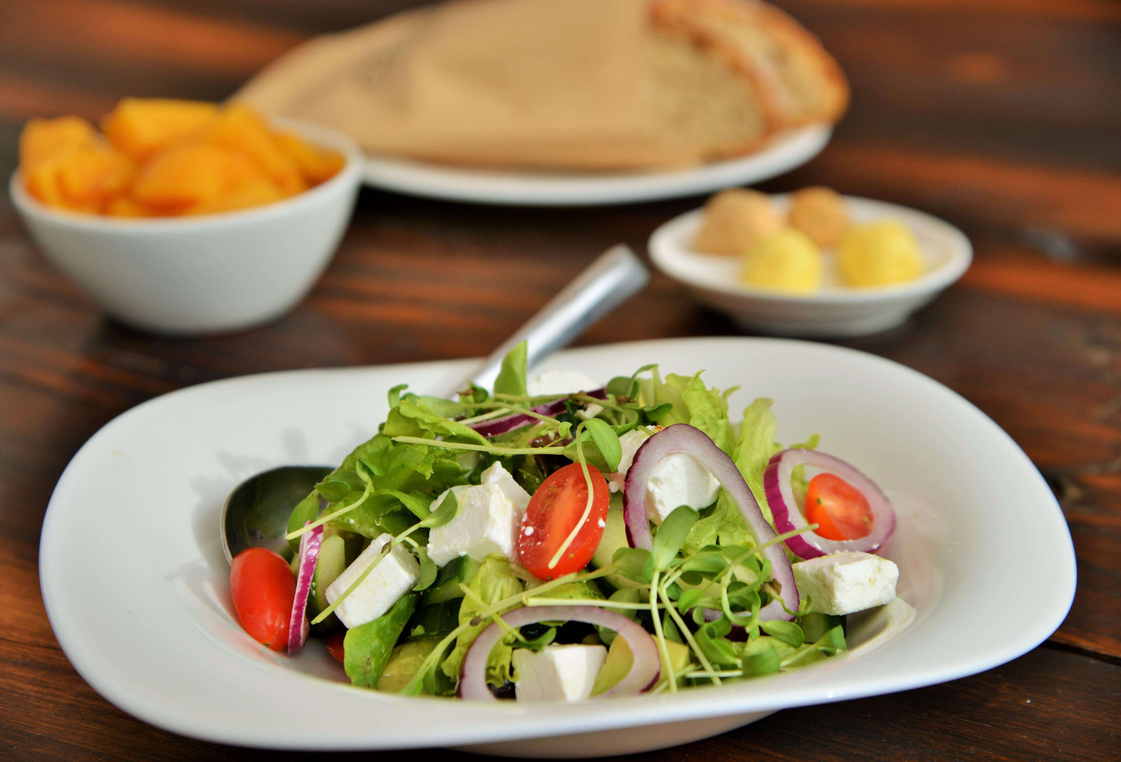 A white plate with a salad, a bowl of fruit, and a plate of bread on a wooden table.