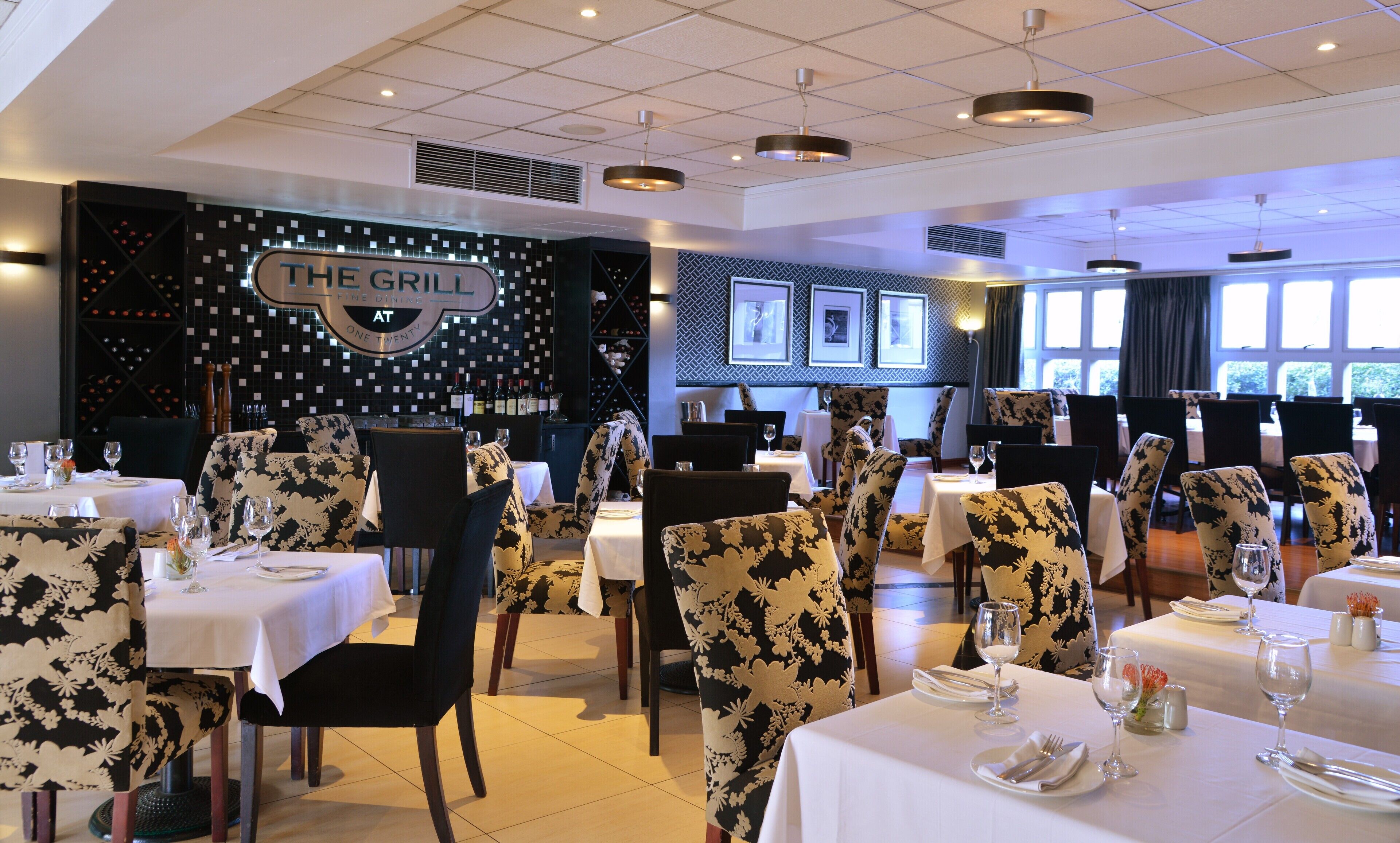 A restaurant interior with tables set for dining, chairs with floral upholstery, and a wall with framed pictures.