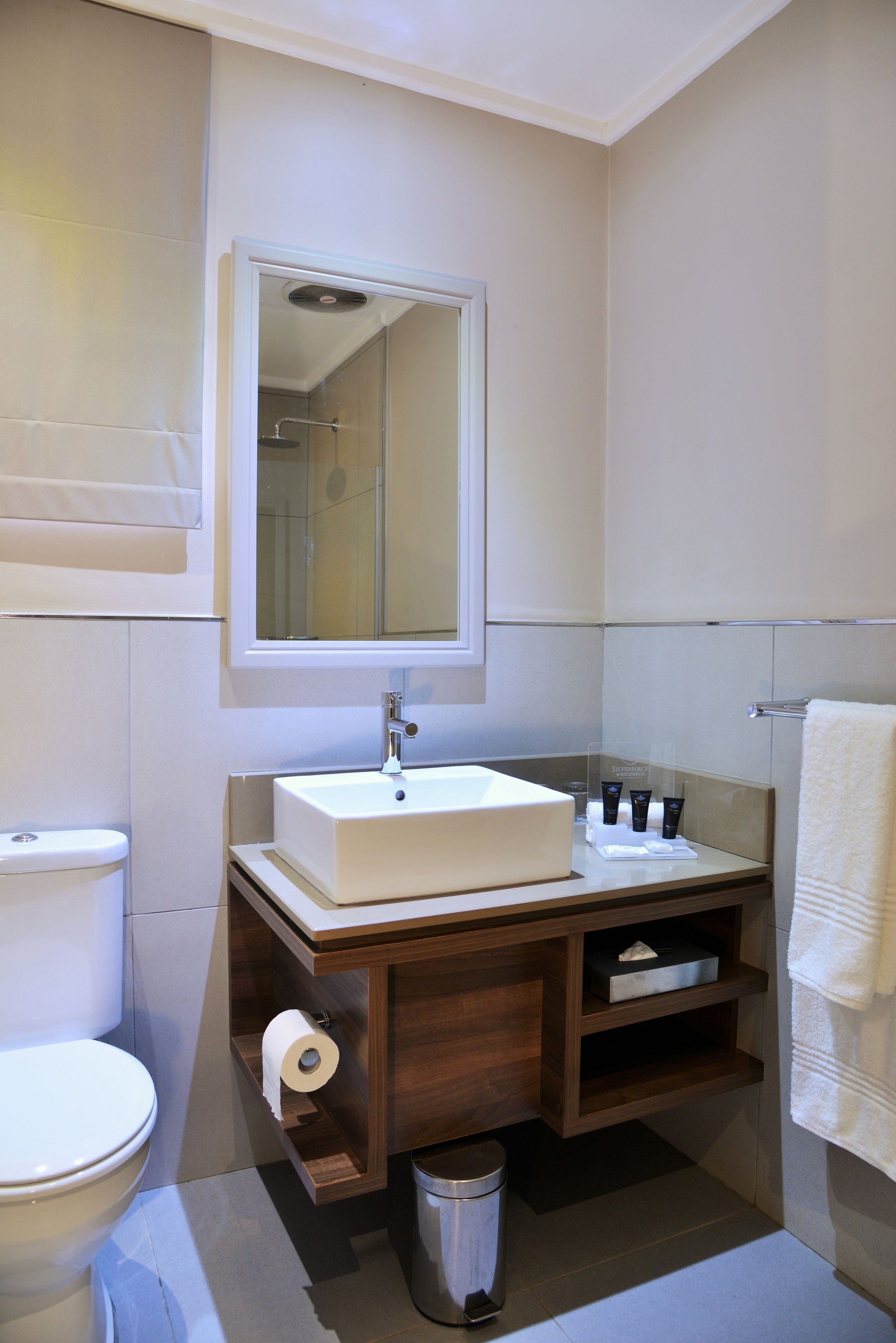 A bathroom with a white sink, mirror, and toilet. There is a towel rack and a trash can.