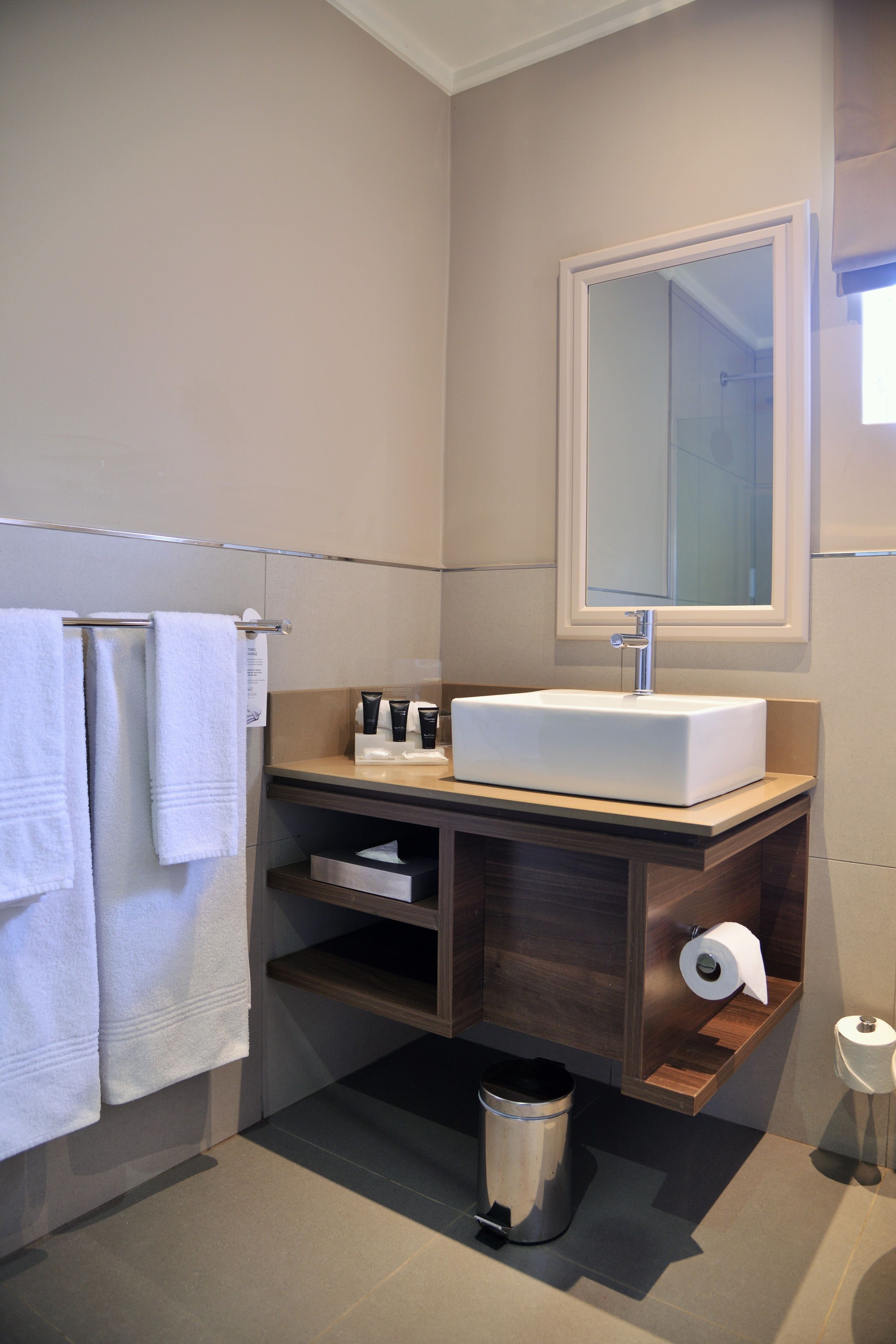 A modern bathroom with a white sink, a mirror, a towel rack, a trash bin, and a toilet paper holder.