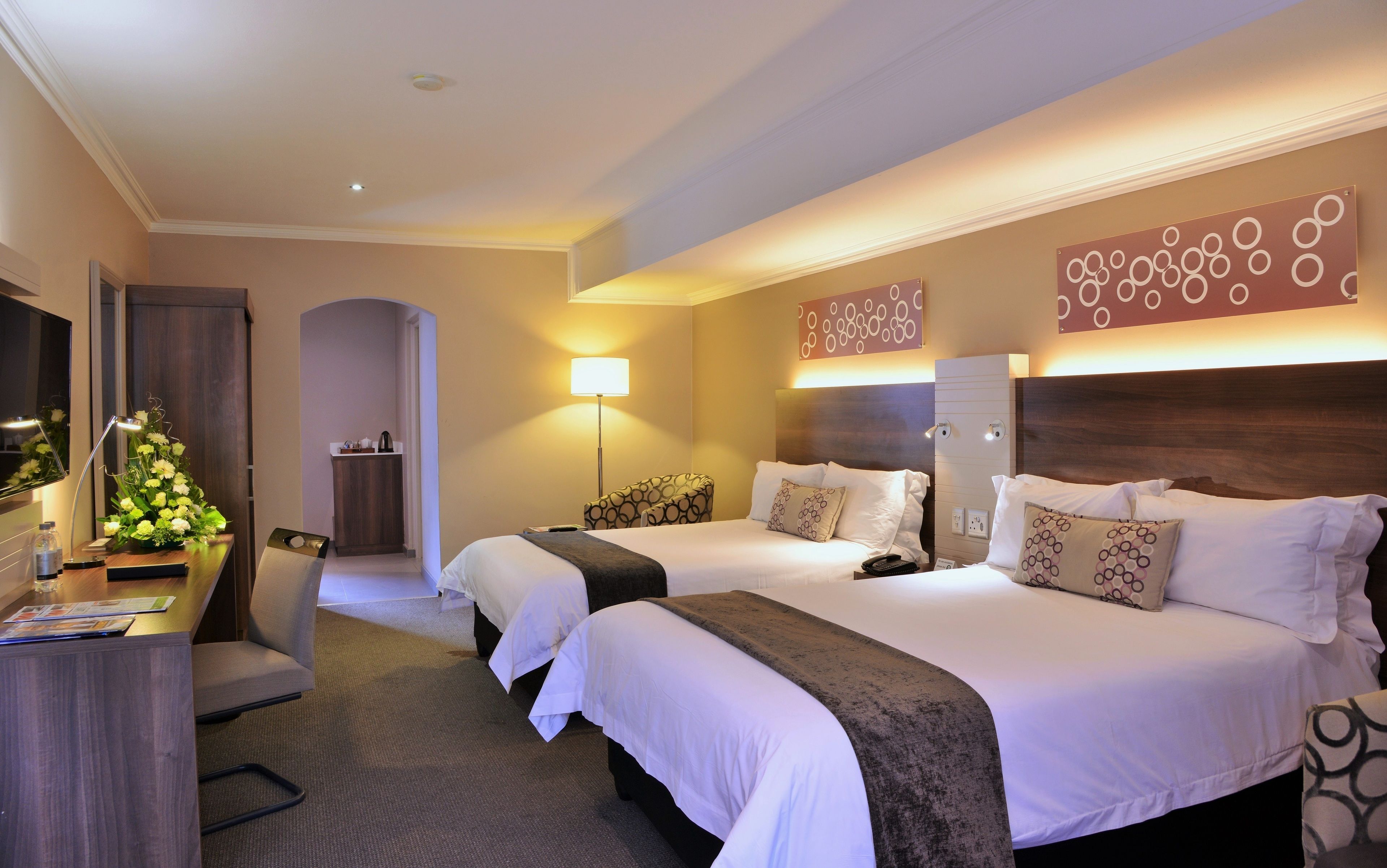 A hotel room with two beds, a desk, a chair, a lamp, and a decorative wall piece.