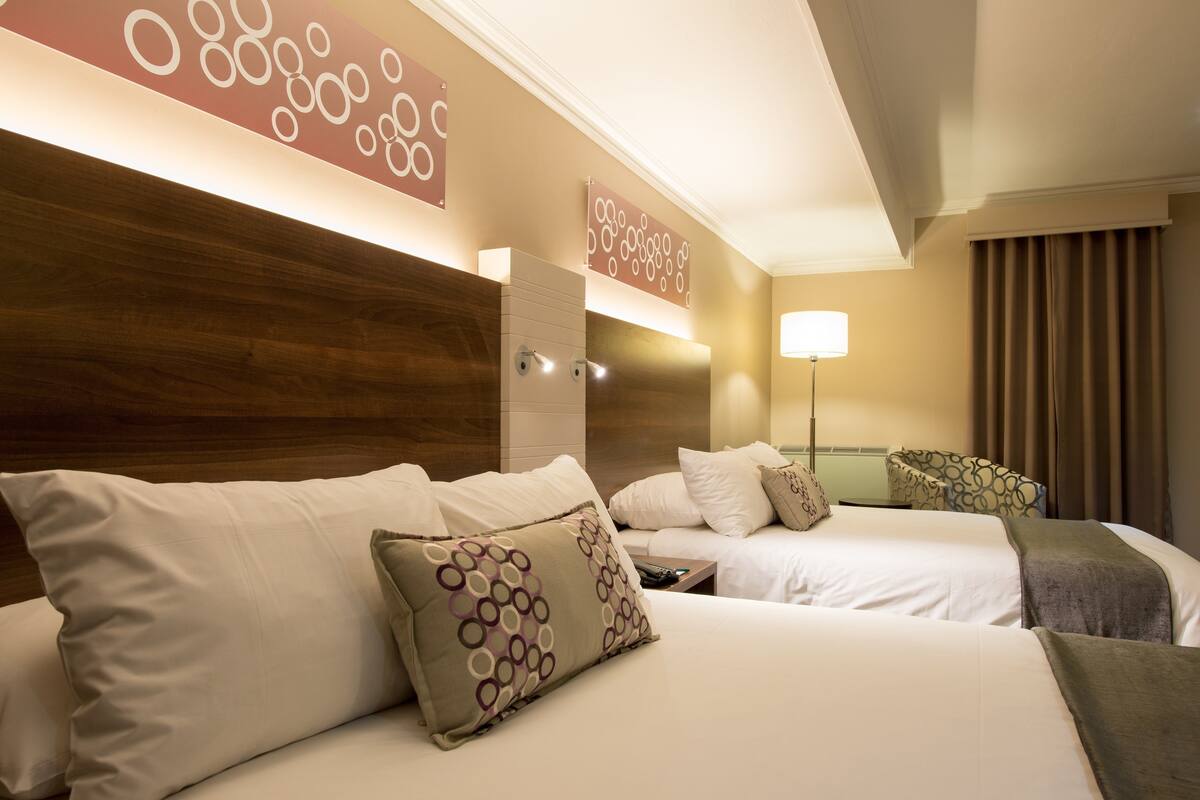 Habitación de hotel con dos camas, cabecero de madera, lámpara de pared y un ventanal con cortinas.