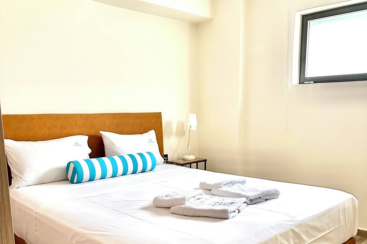 Un lit bien fait, avec des draps blancs, un coussin à rayures et des serviettes pliées, posé sur un plancher en bois.