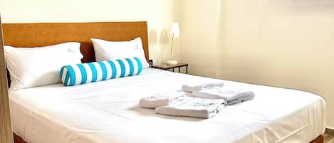 A neatly made bed with white linens, a striped pillow, and folded towels on a wooden floor.