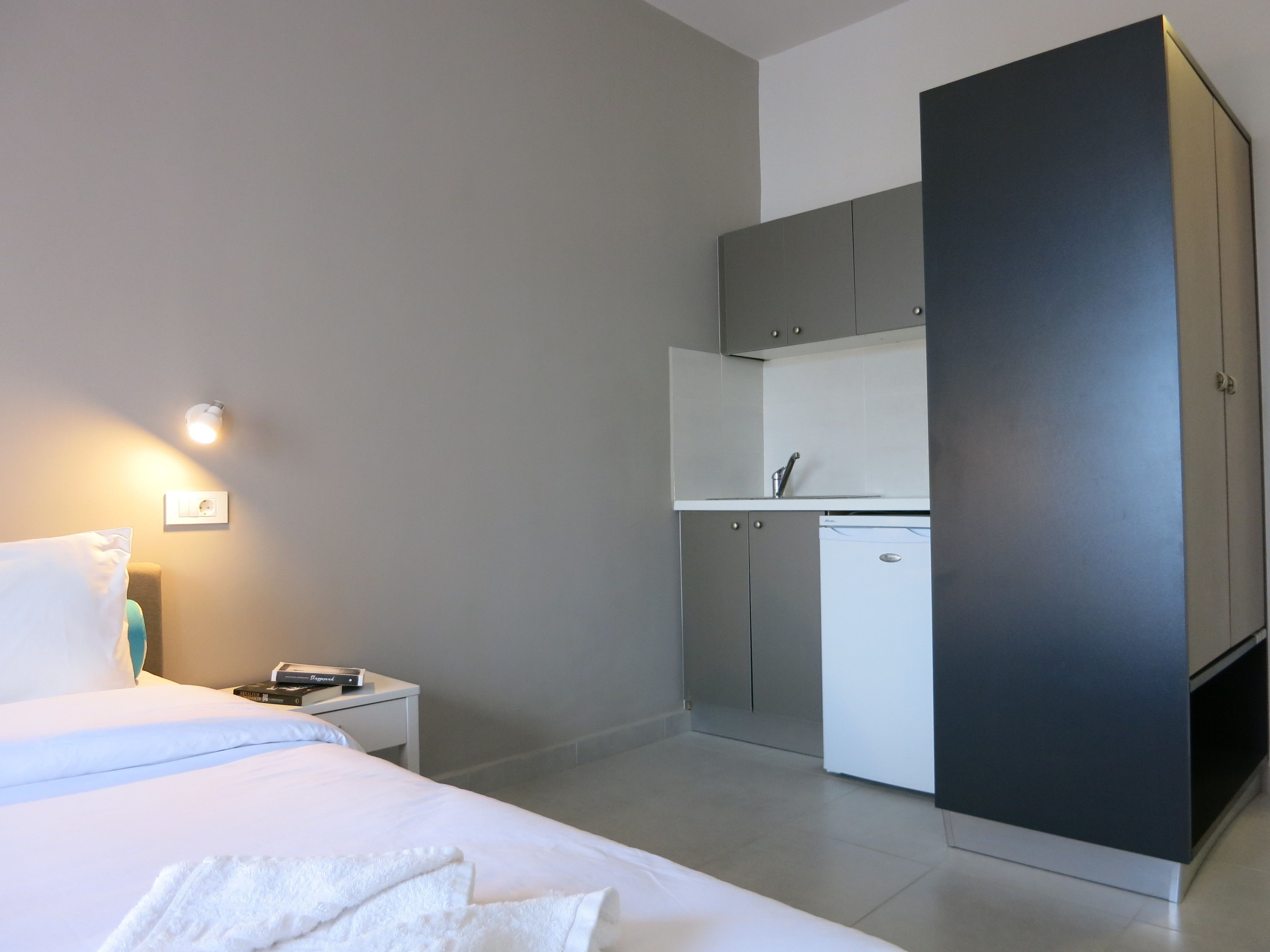 A modern hotel room with a bed, bedside table, and a kitchenette area.
