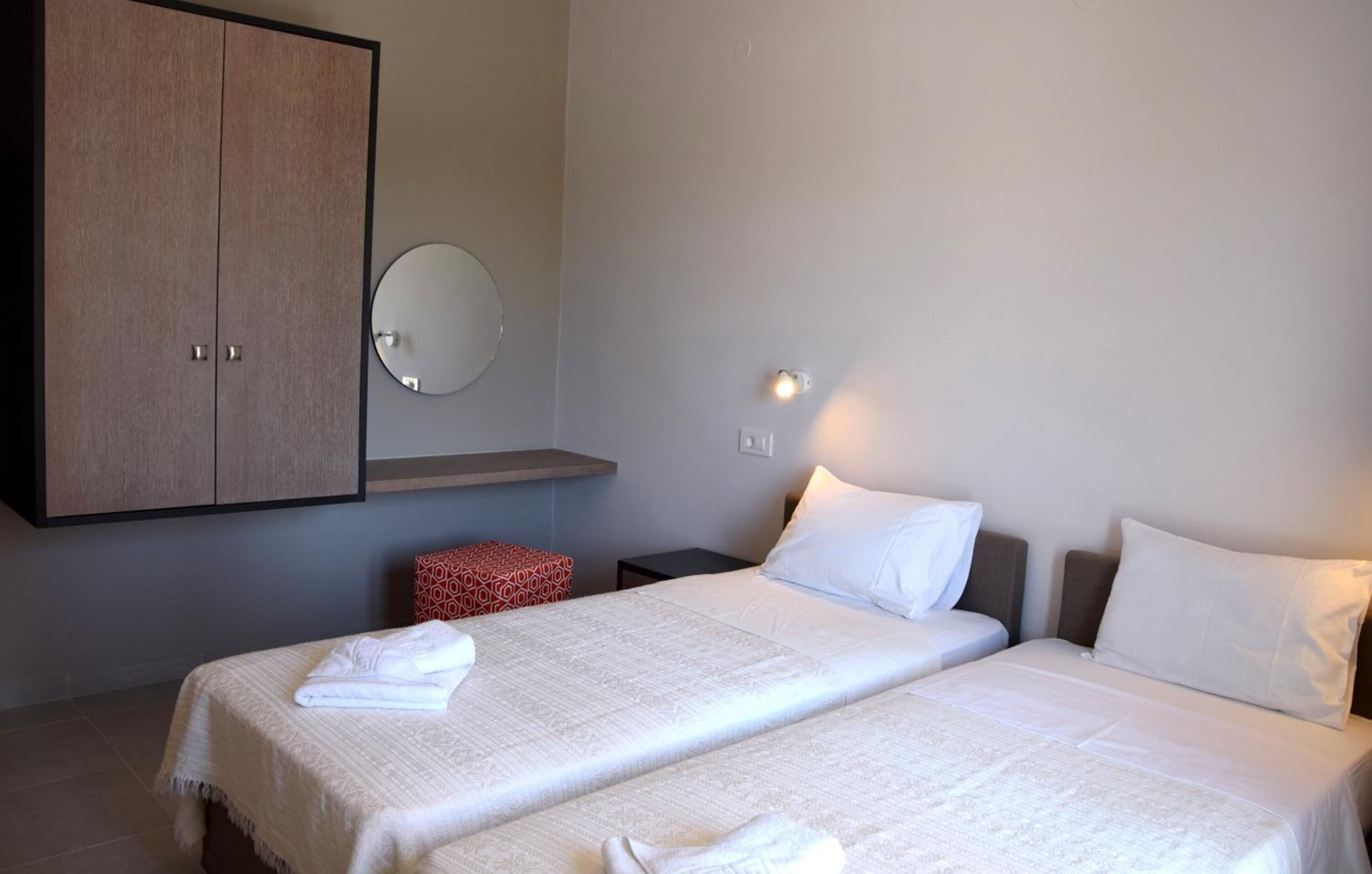 Habitación de hotel con dos camas, un armario, un espejo redondo y un pequeño taburete rojo.