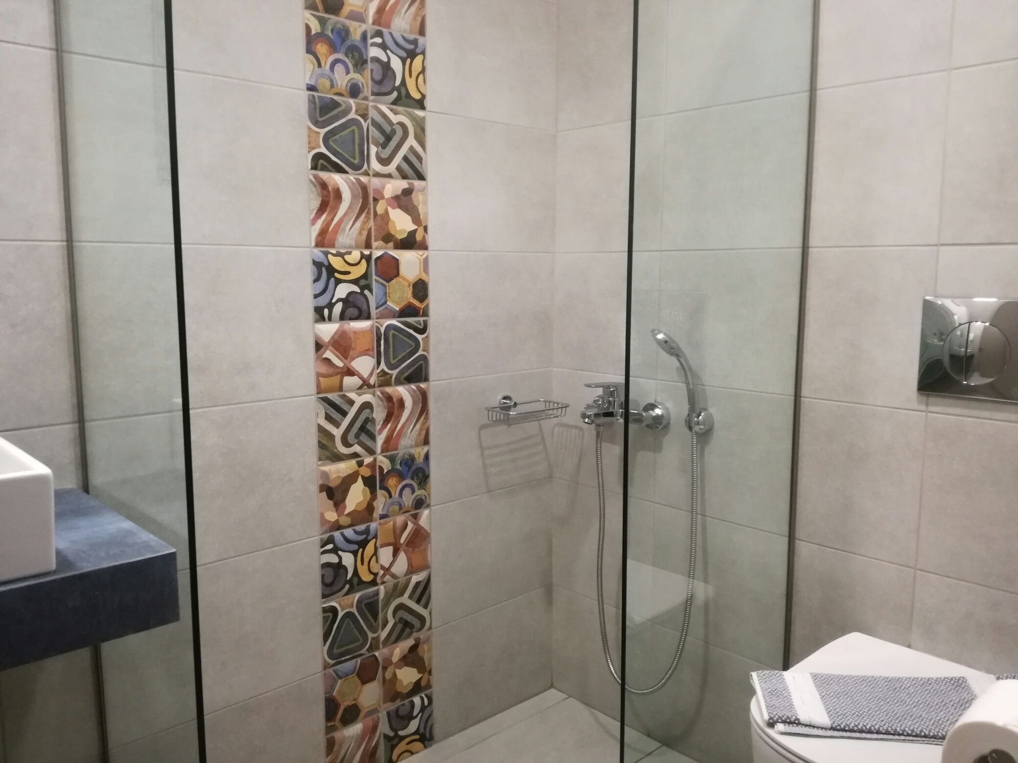 A shower area with a glass door, a tiled wall with a colorful pattern, and a toilet with a towel and a tissue box.