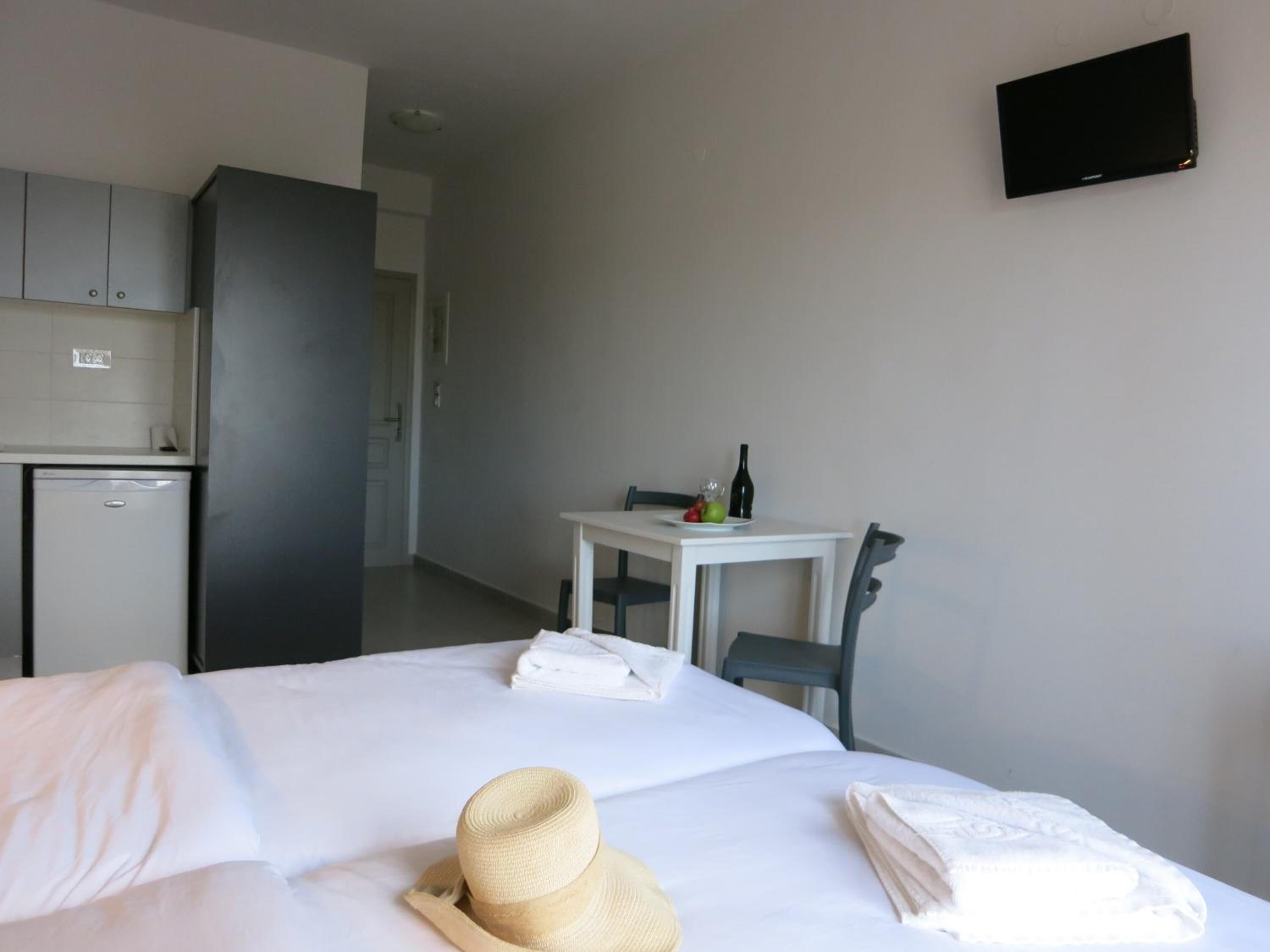 A hotel room with a bed, a small table with a bottle and fruit, a TV on the wall, and a kitchenette in the background.
