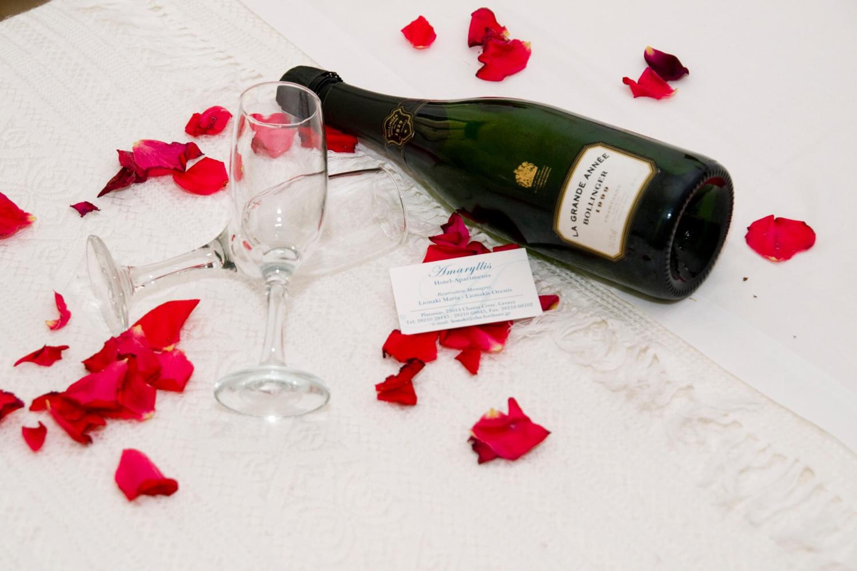A bottle of wine, a glass, and a card on a white table with red rose petals.