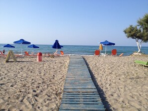 A sandy beach with a wooden walkway leading to the sea, surrounded by red and blue beach chairs and umbrellas. - Baladinos Apartments  (Platanias)