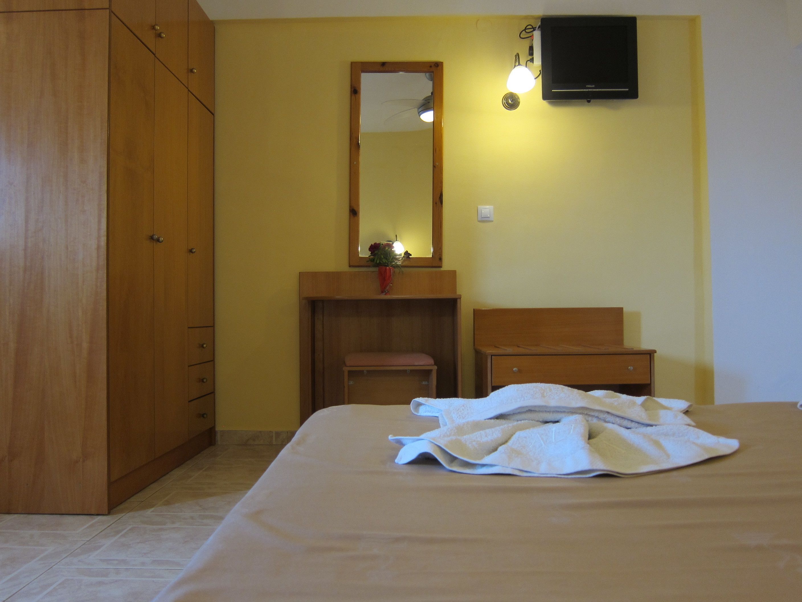 Une chambre avec une armoire en bois, une table de chevet, un miroir et un lit recouvert d’une couverture blanche.