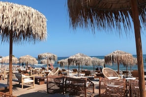 Eine Strandlounge mit Reetdachschirmen, Korbsesseln und Holztischen.