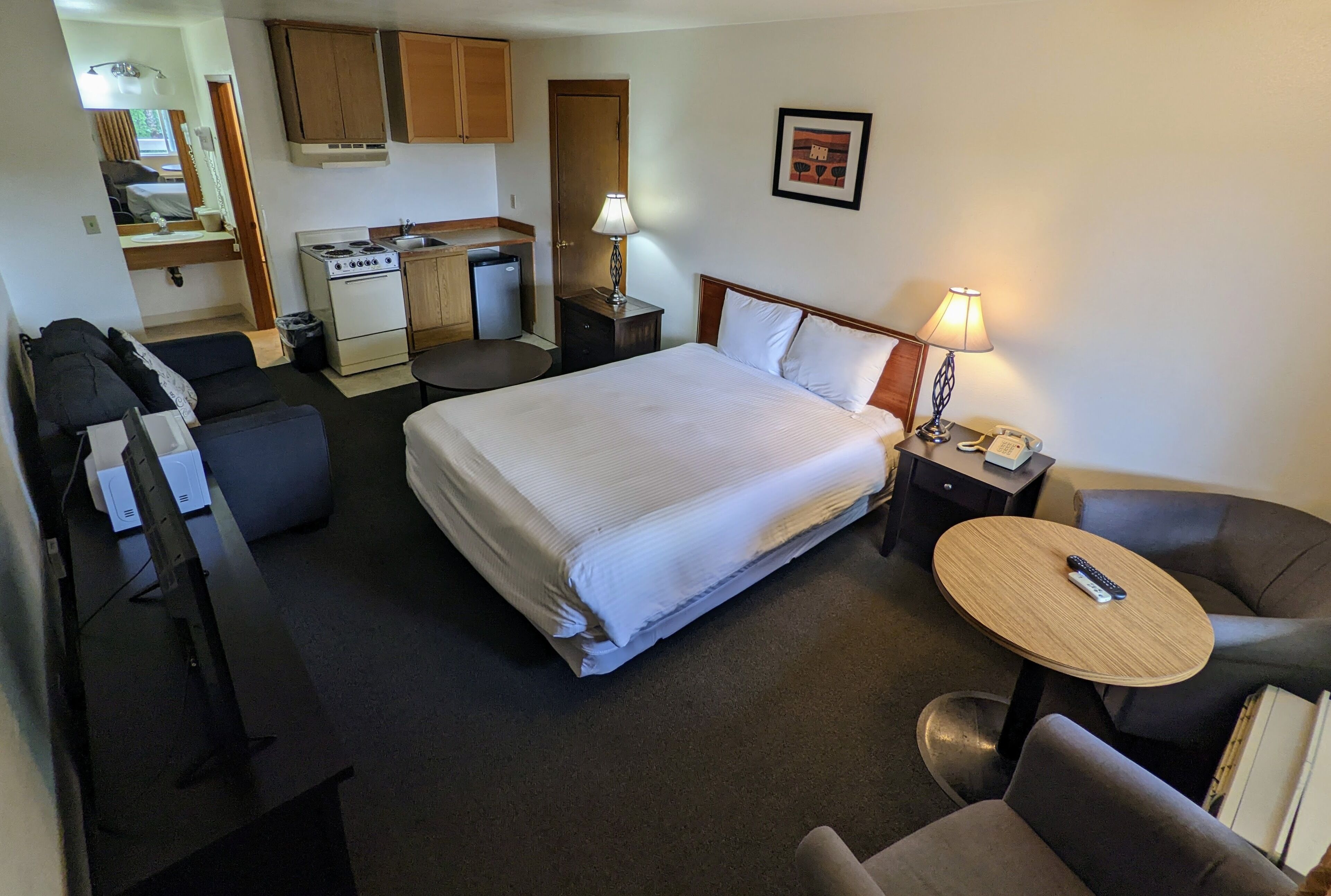 A hotel room with a large bed, a desk with a computer, a chair, a small table with a lamp, a kitchenette, and a bathroom.