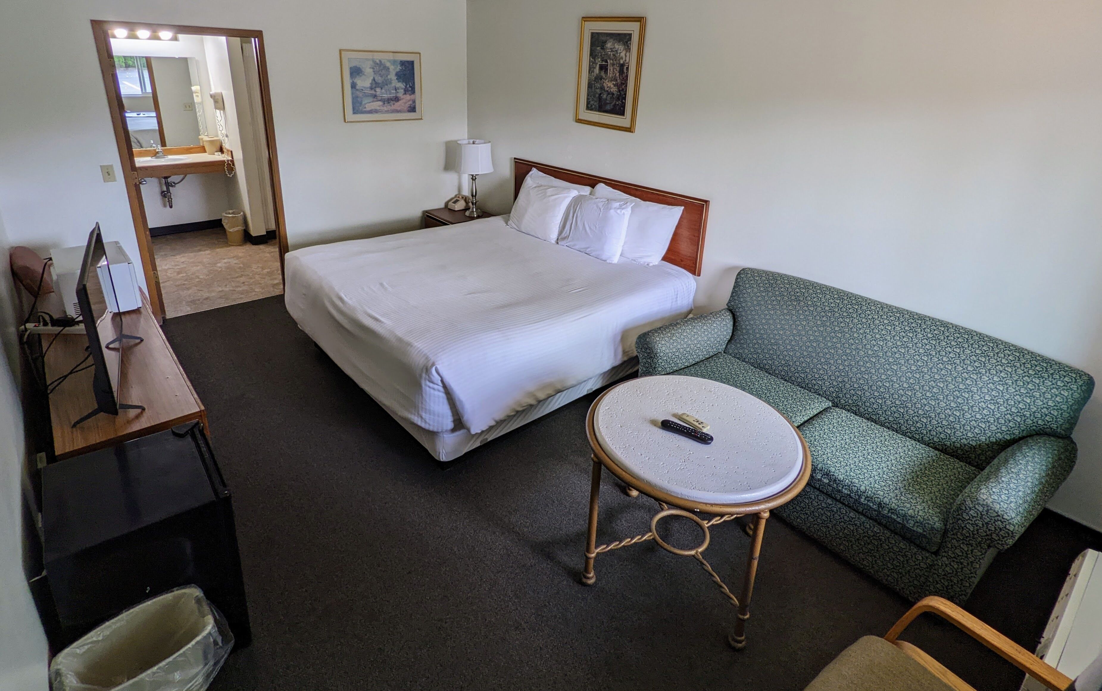 A hotel room with a bed, a green patterned sofa, a small round table with a remote control, a desk with a computer, and a mirror.