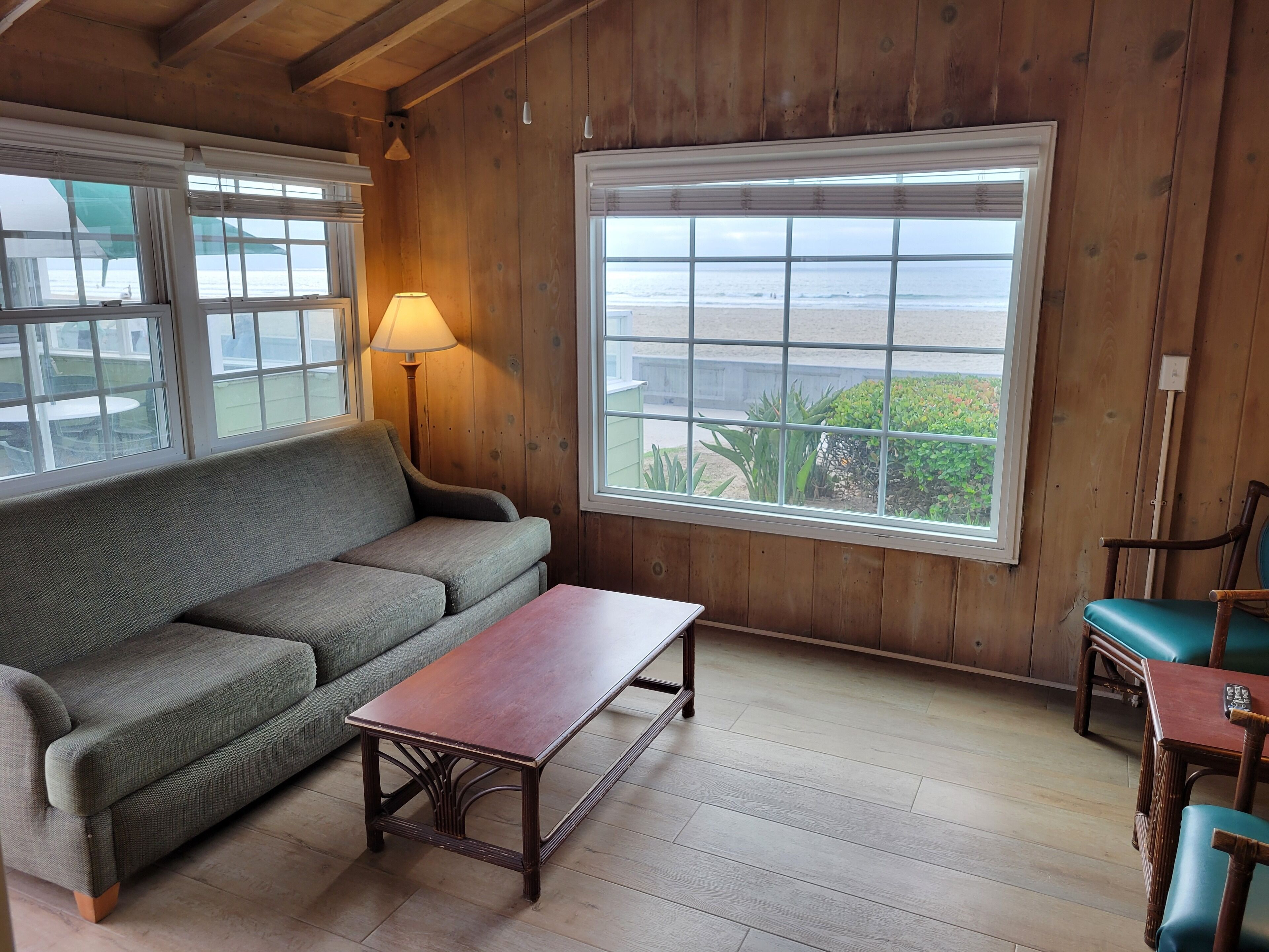 A cozy room with a wooden ceiling, a green sofa, a wooden table, and a large window with a view of the beach.