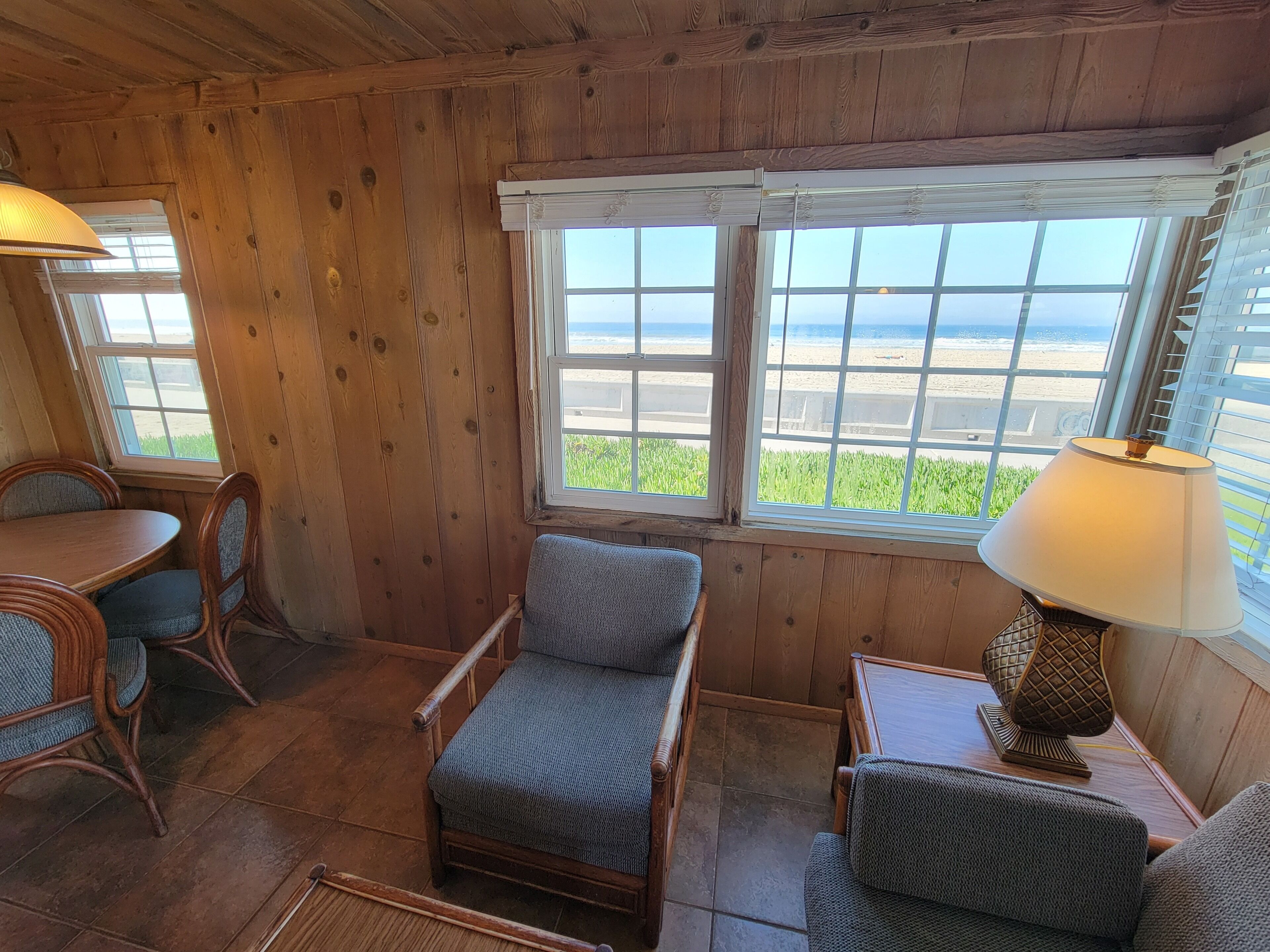 A wooden cabin interior with a dining table, two chairs, a cushioned chair, a lamp, and a window with a beach view.