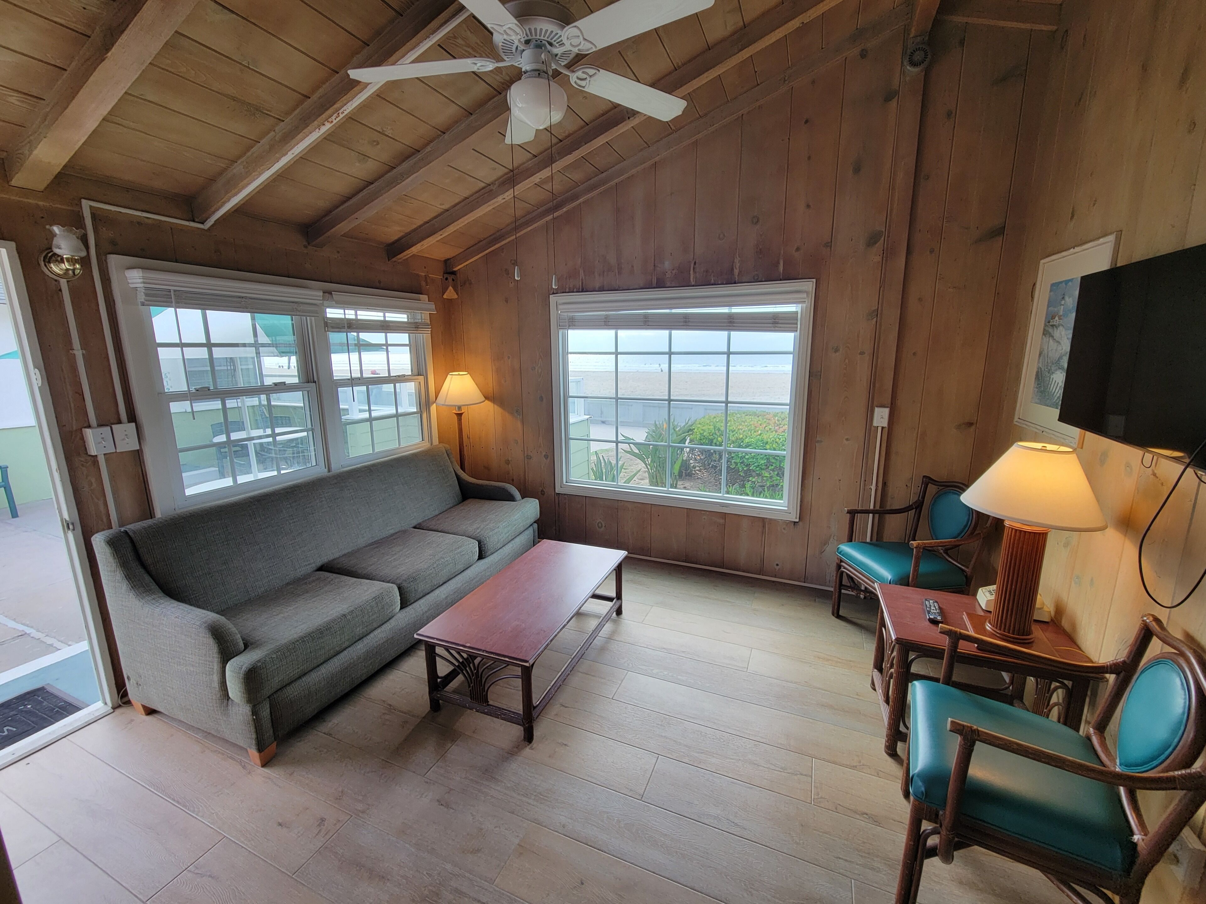 Ein Wohnzimmer mit Holzboden, einem Sofa, einem Couchtisch und zwei Sesseln. Große Fenster bieten einen Ausblick ins Freie.