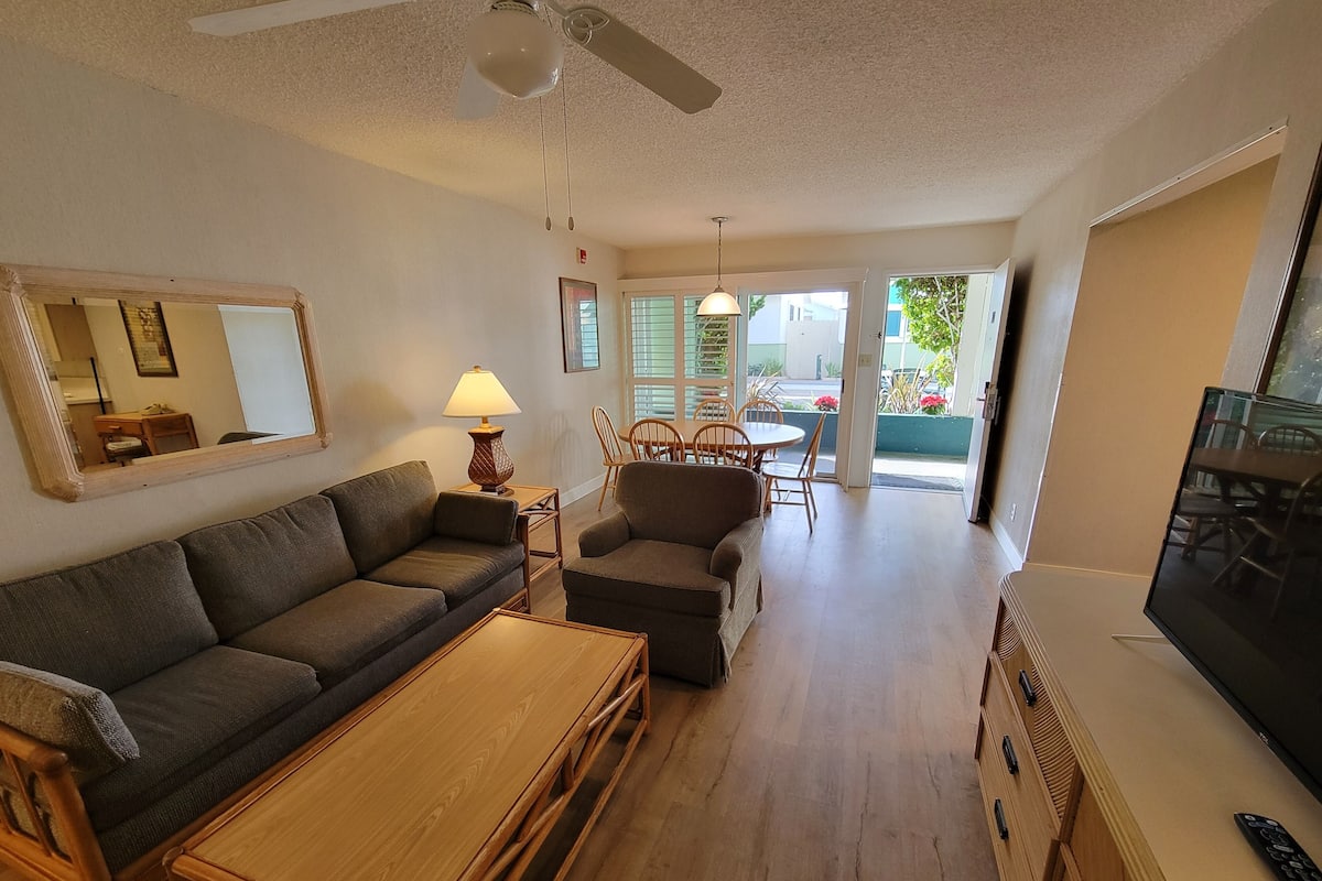 Ett vardagsrum med en soffa, ett träbord, en lampa och en tv. I bakgrunden finns en matplats med stolar och ett bord.