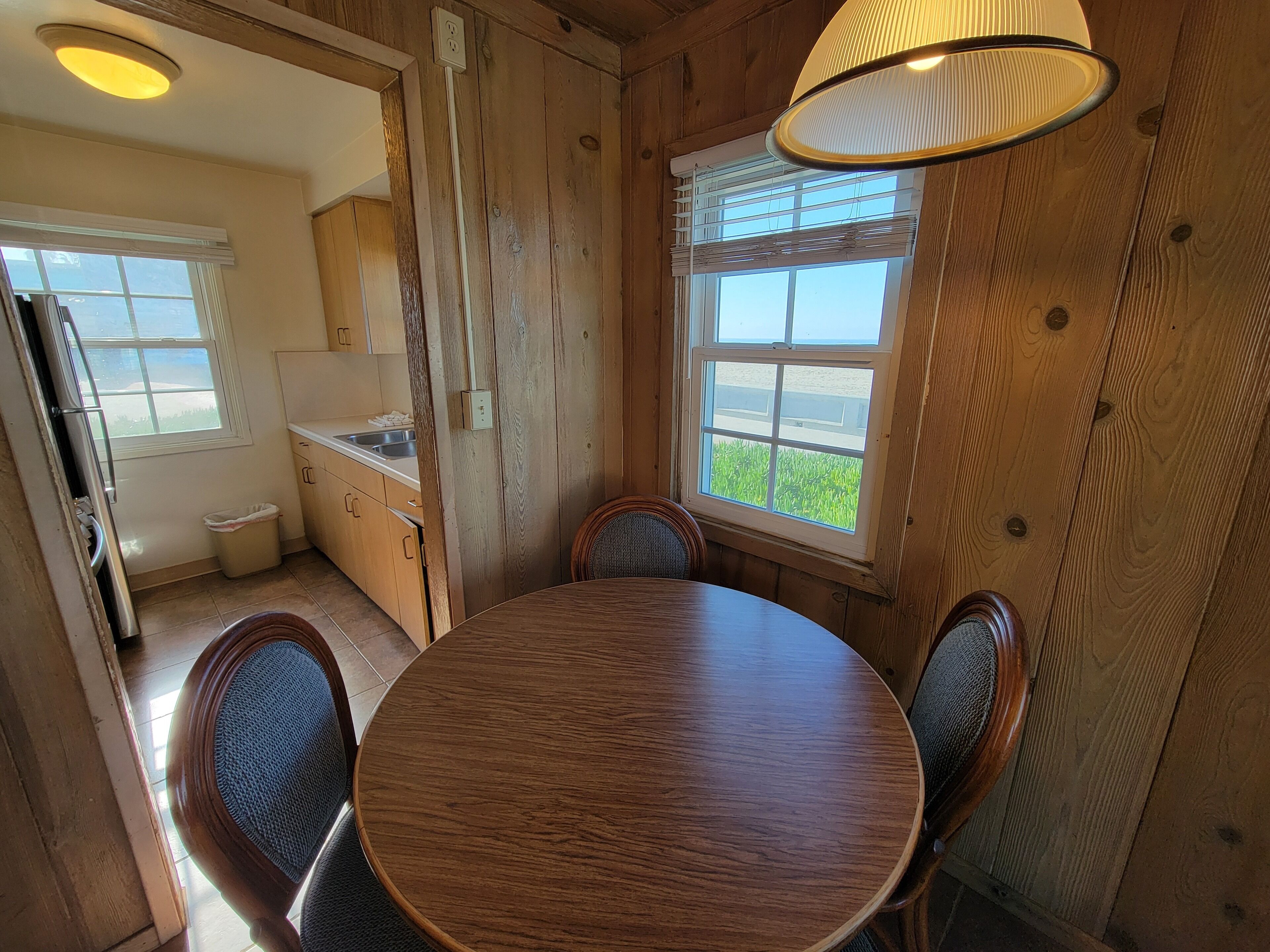 A wooden interior with a round dining table and chairs, a window with blinds, and a ceiling light fixture.