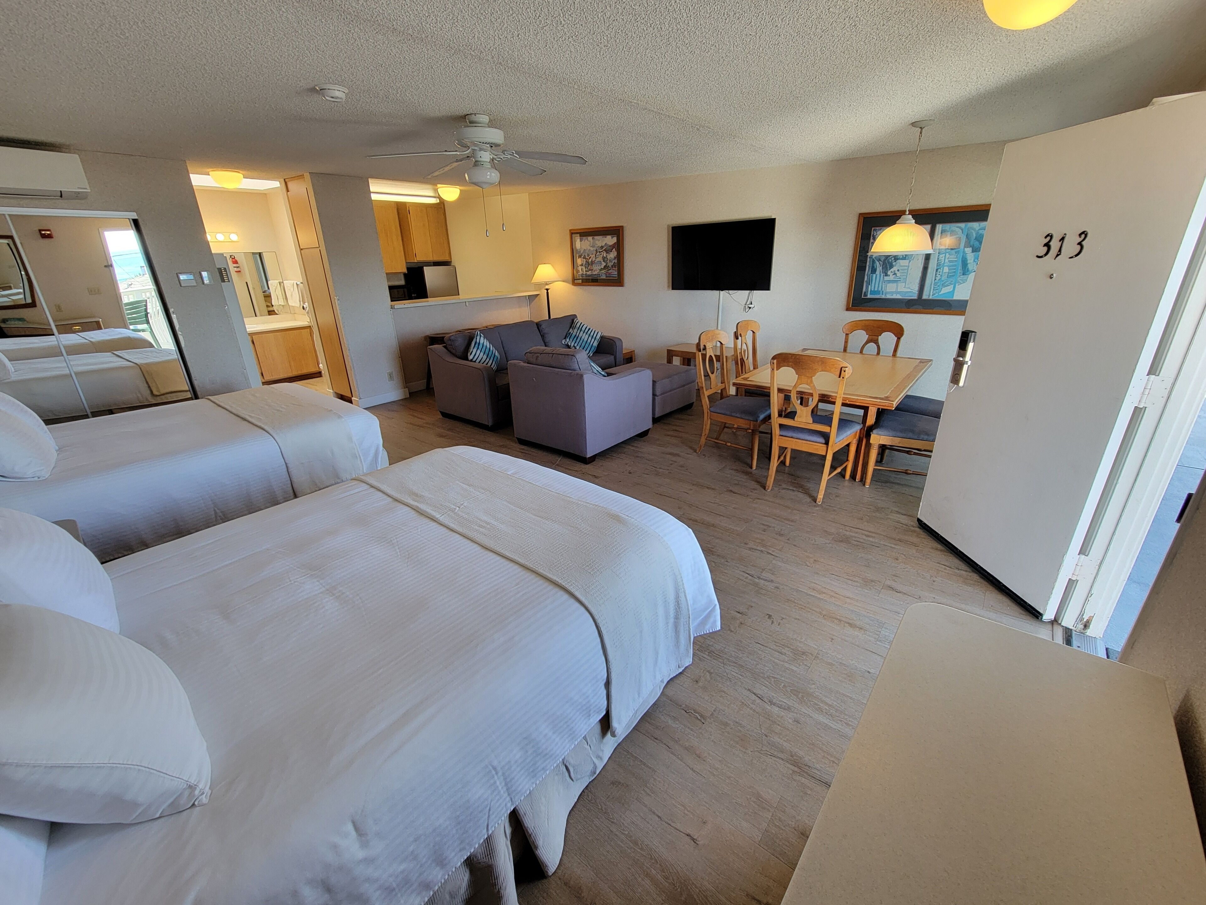 Habitación de hotel con dos camas, un sofá, una mesa de comedor con sillas, un ventilador de techo, un televisor y un refrigerador.