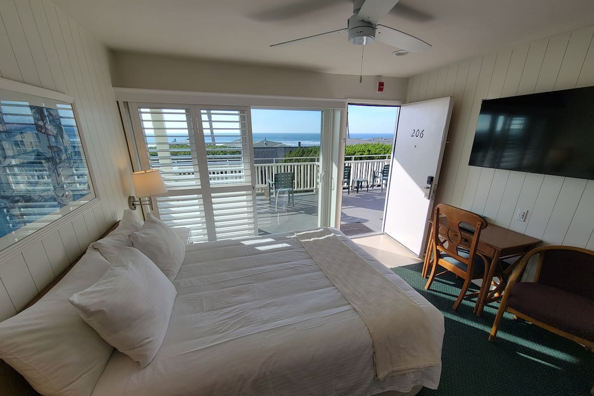 Ett hotellrum med en säng, ett skrivbord, en stol, en TV och en balkong med utsikt över havet.