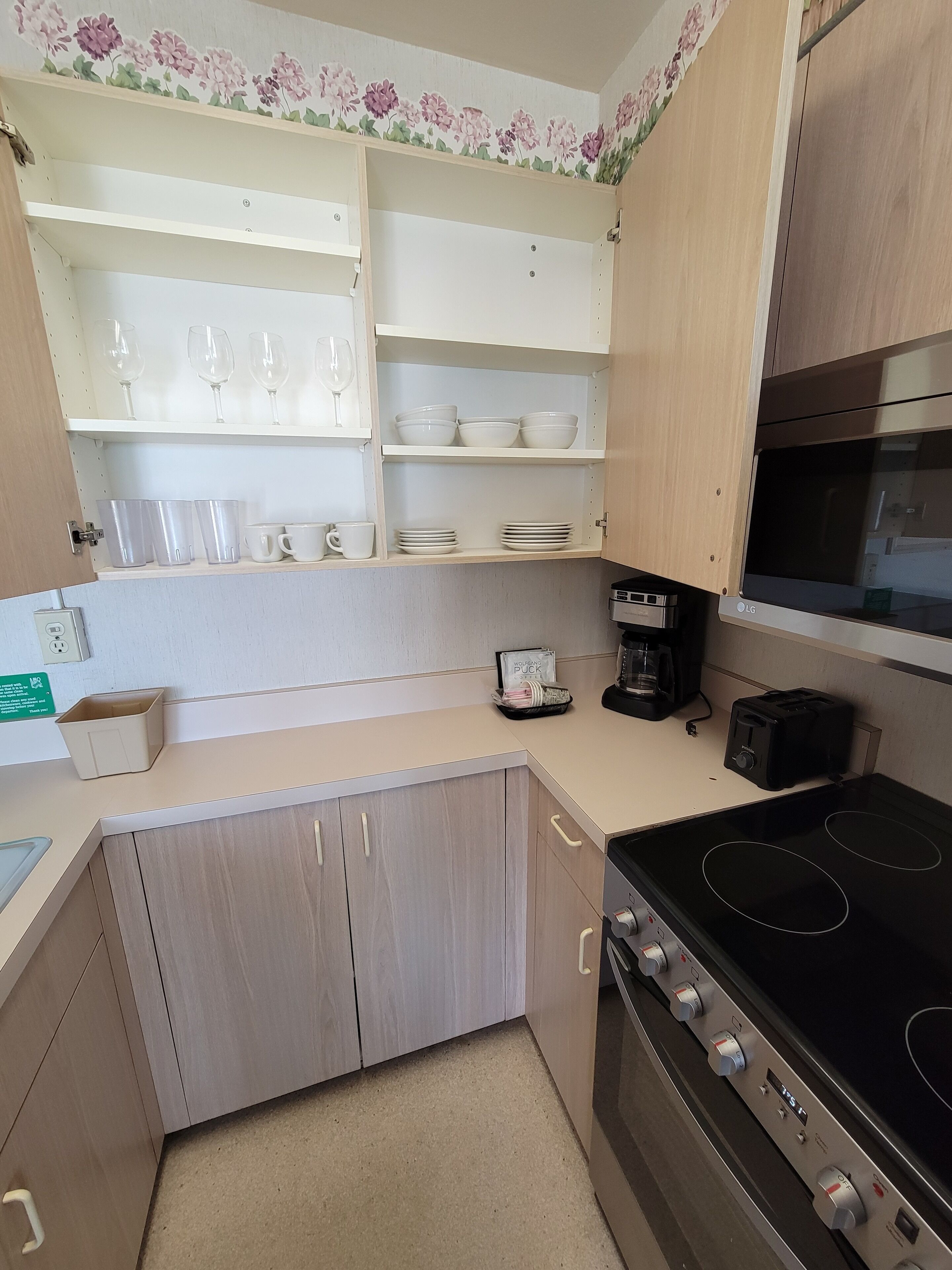 Uma cozinha compacta com fogão, micro-ondas e cafeteira. Há armários com prateleiras acima para guardar pratos e copos.