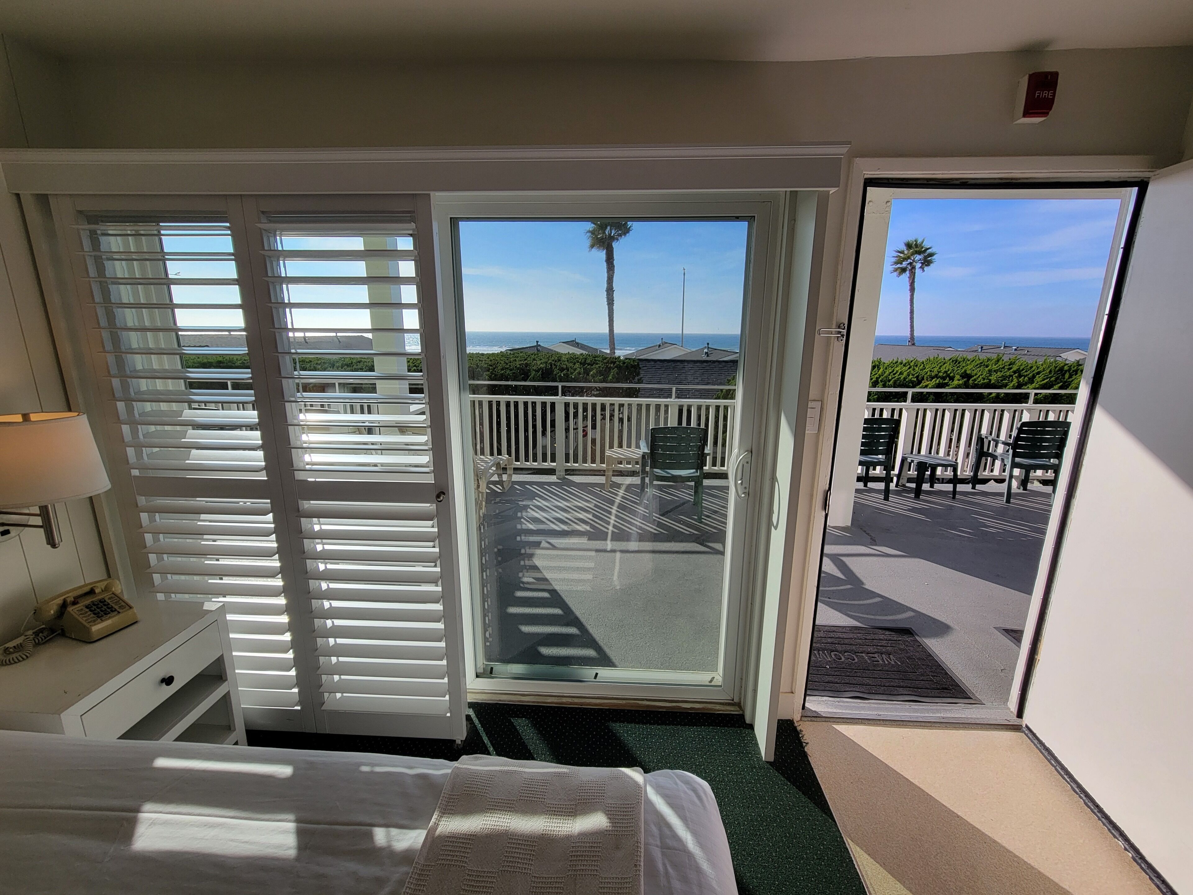 A hotel room with a bed, a desk with a lamp, a sliding glass door leading to a balcony with chairs and a view of the ocean.